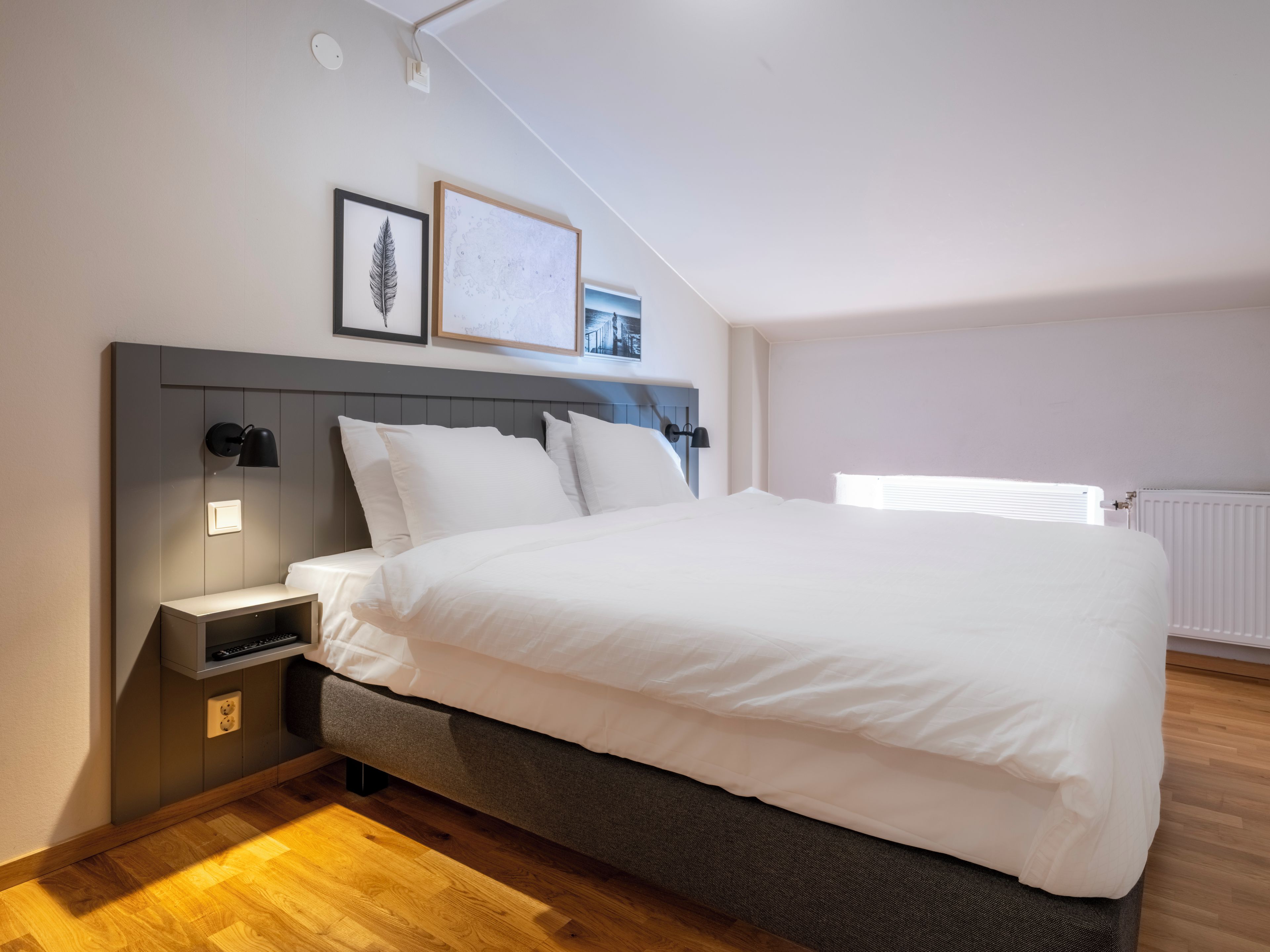 A modern bedroom with a large bed, a bedside table, a wall-mounted lamp, and a framed picture.