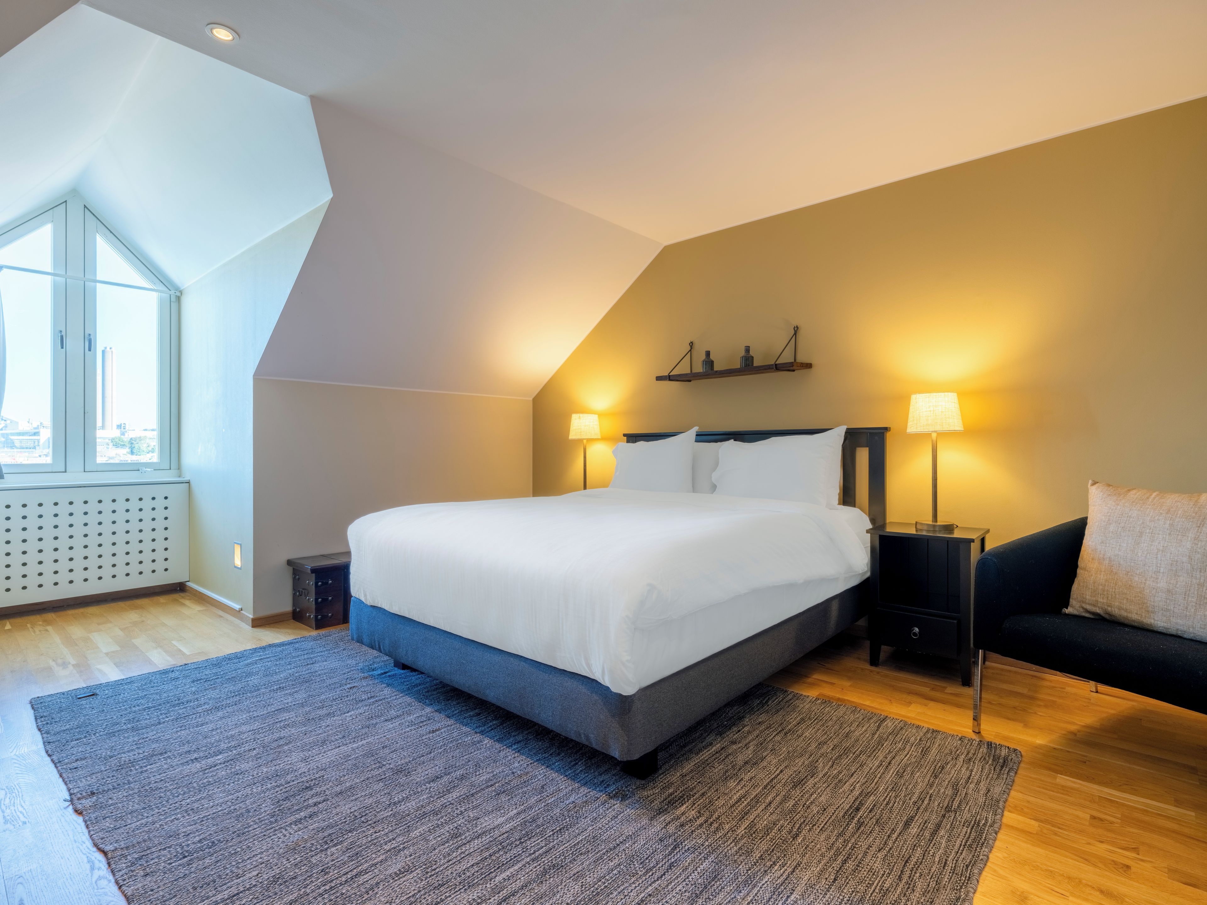 A modern bedroom with a large bed, two bedside lamps, a dark sofa, and a view of the city through a large window.