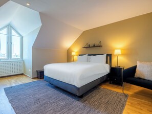 A modern bedroom with a large bed, two bedside lamps, a dark sofa, and a view of the city through a large window. - Mercure Gothenburg (Vastra Frolunda)