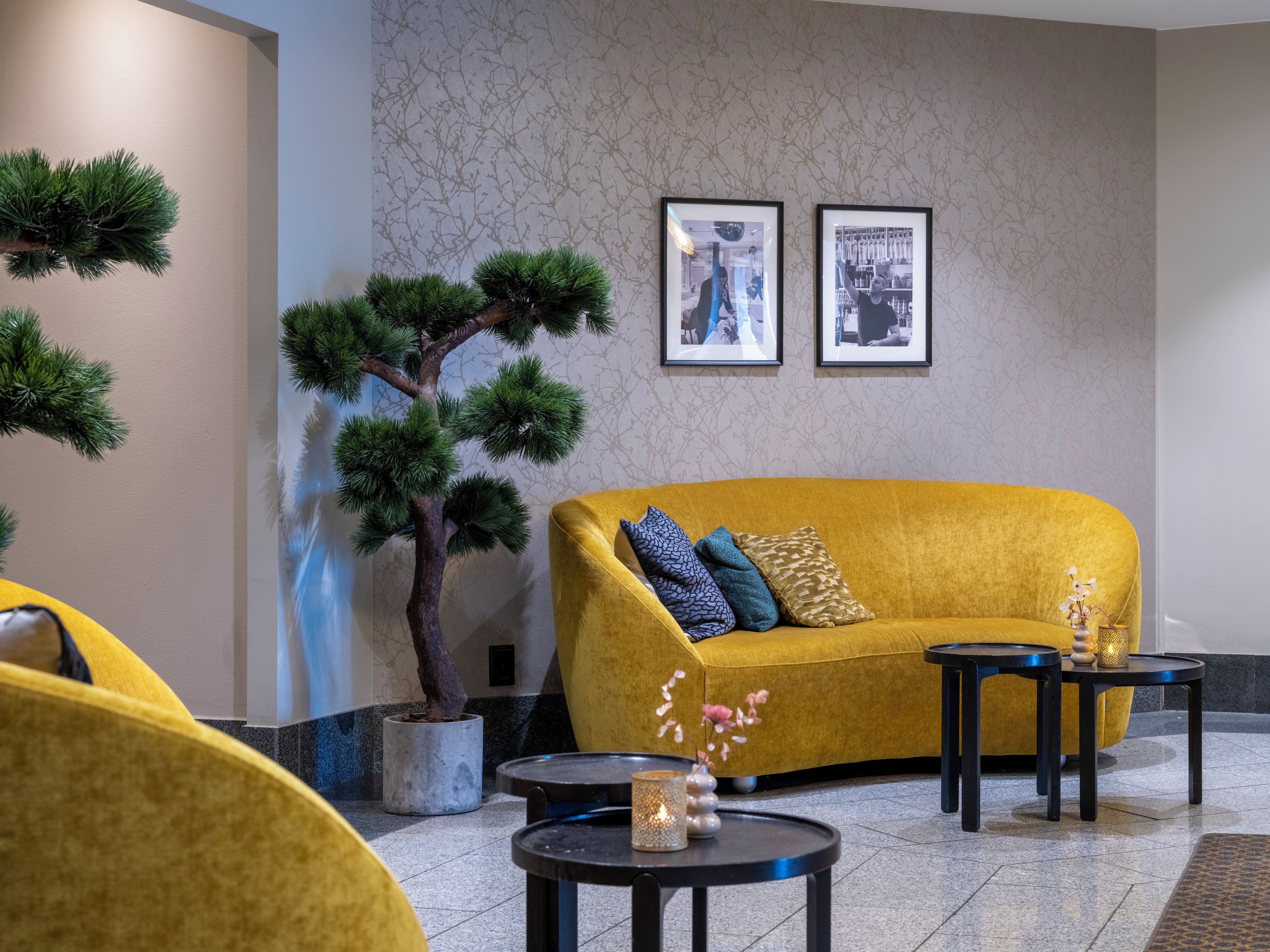 A modern interior with a yellow sofa, a potted tree, framed pictures, and a small table with a candle.