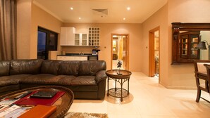 A living room with a leather sofa, a coffee table with remote controls, a small round table, and a kitchen in the background. - Victoria Crown Plaza Hotel (Lagos)