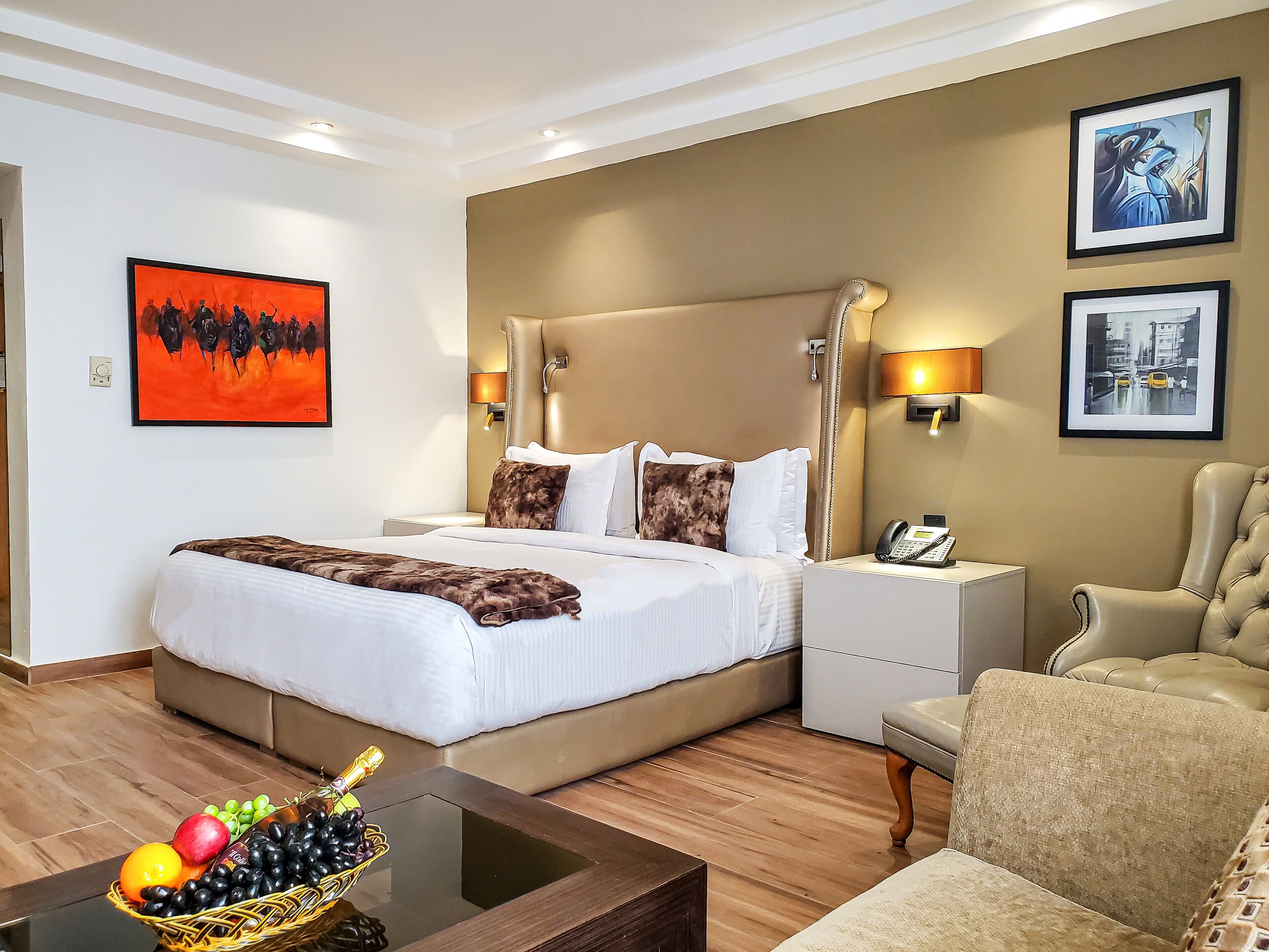 A hotel room with a large bed, a bedside table with a phone, a chair, and a fruit basket on a table.