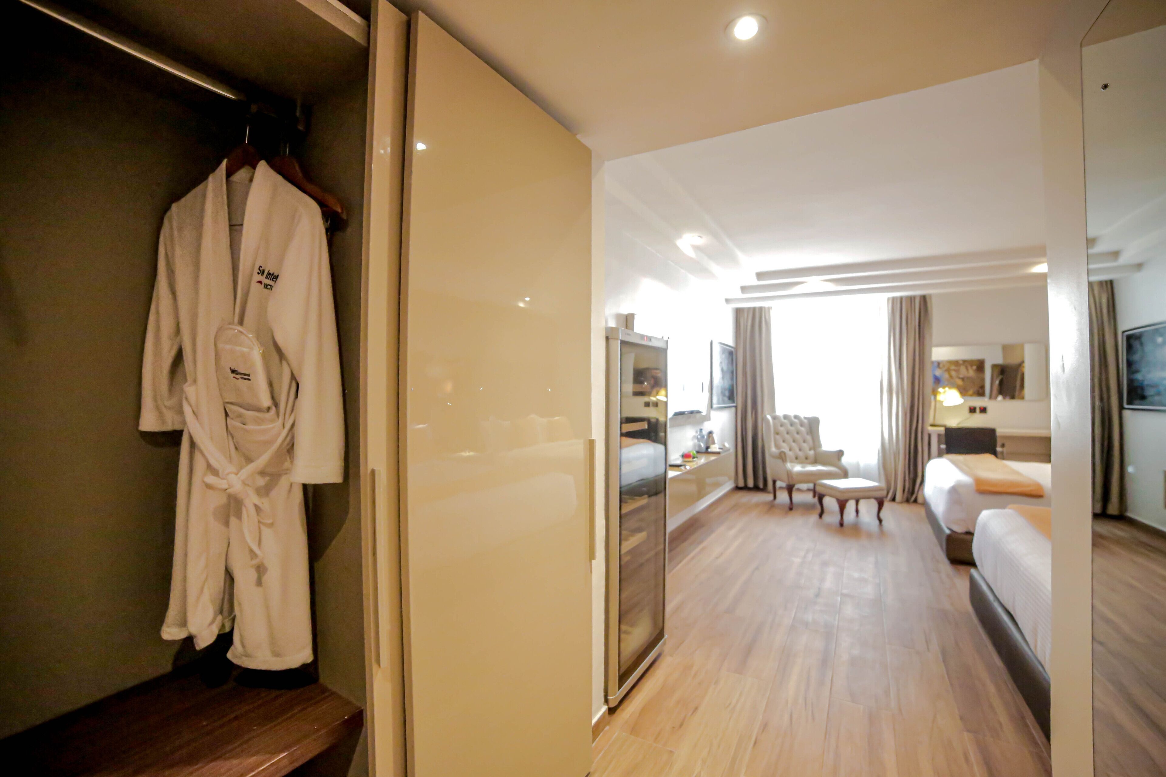 A hotel room with a wooden floor, a bed, a seating area with a chair and ottoman, a robe hanging on a rack, and a mirror.