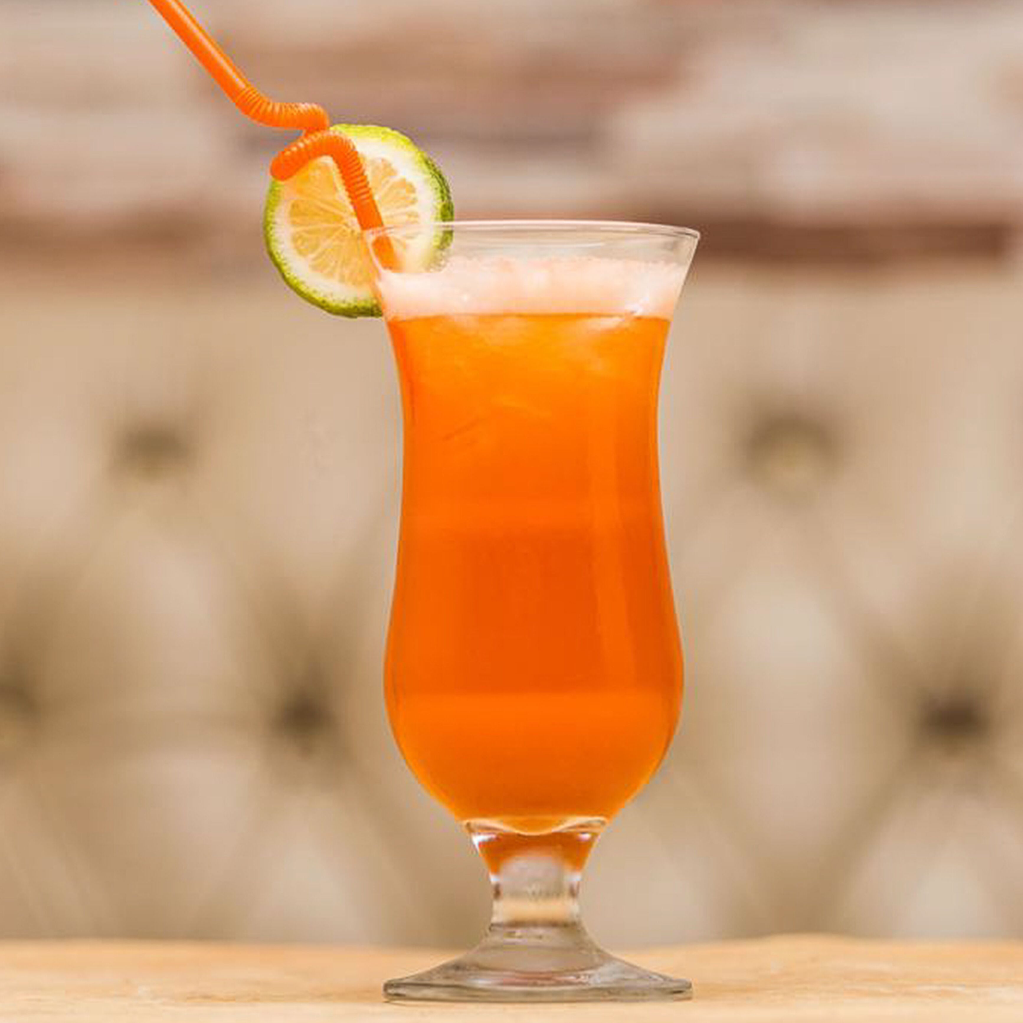 A tall glass with a lime wedge and an orange straw, filled with a bright orange beverage, on a wooden surface.