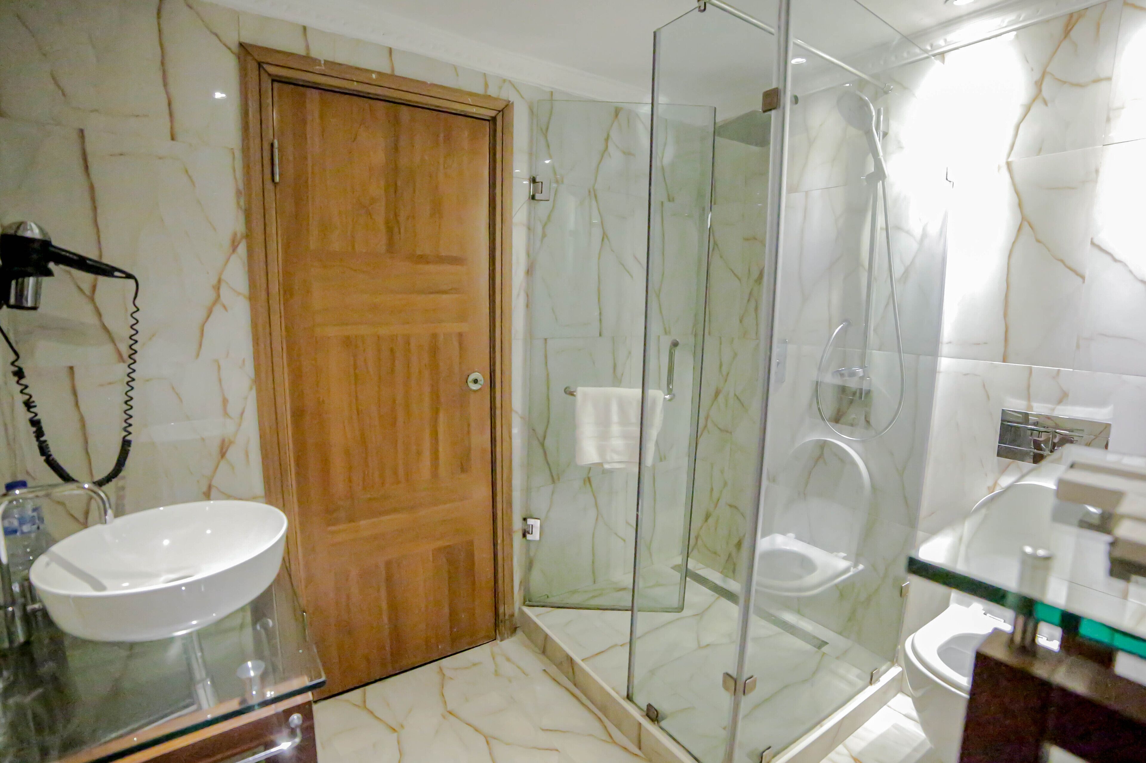 Een badkamer met een houten deur, een douche met glazen wanden, een witte wastafel en een toilet.