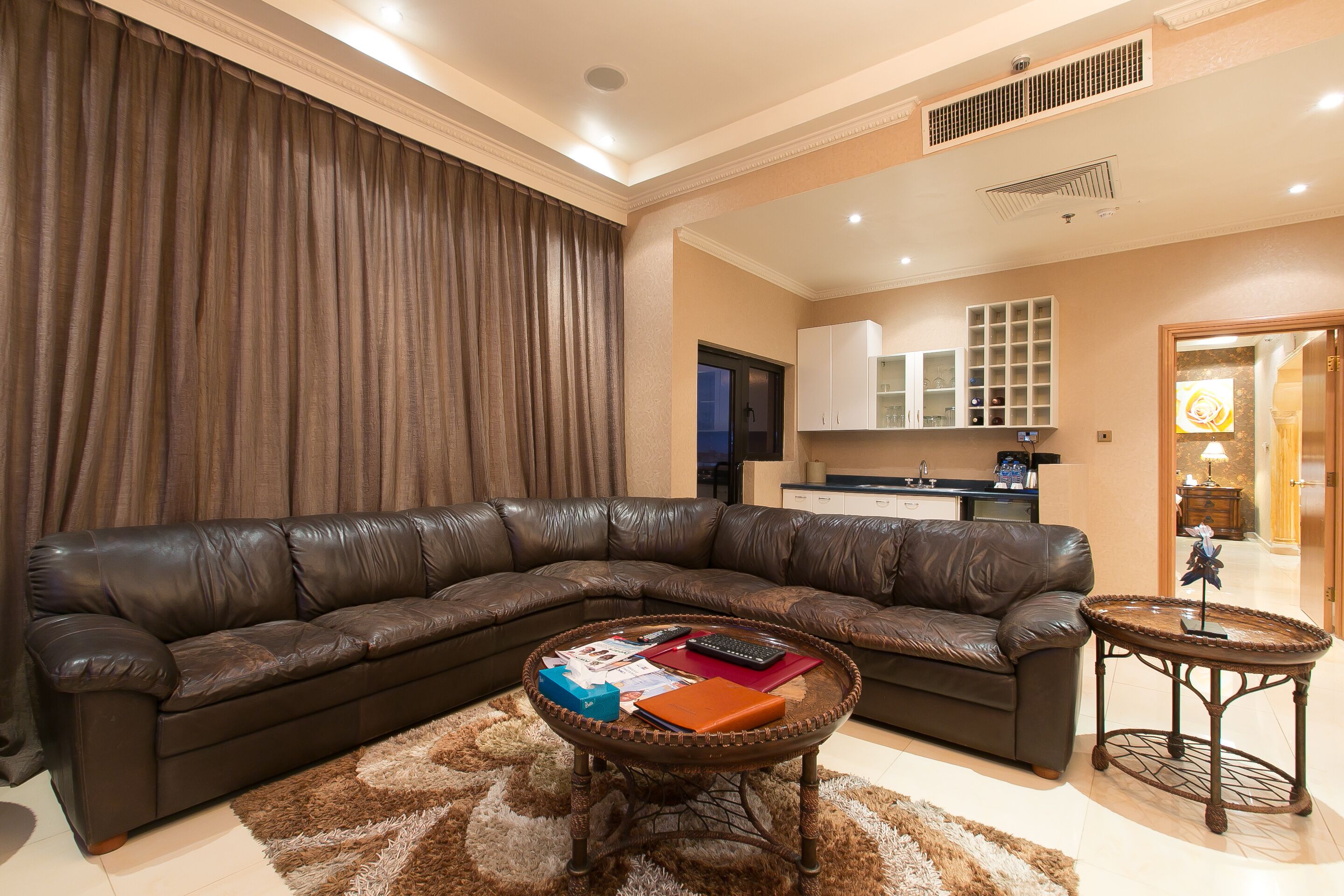A living room with a dark brown leather sectional sofa, a round coffee table with magazines and a remote control, a fluffy brown rug, and a small round side table.