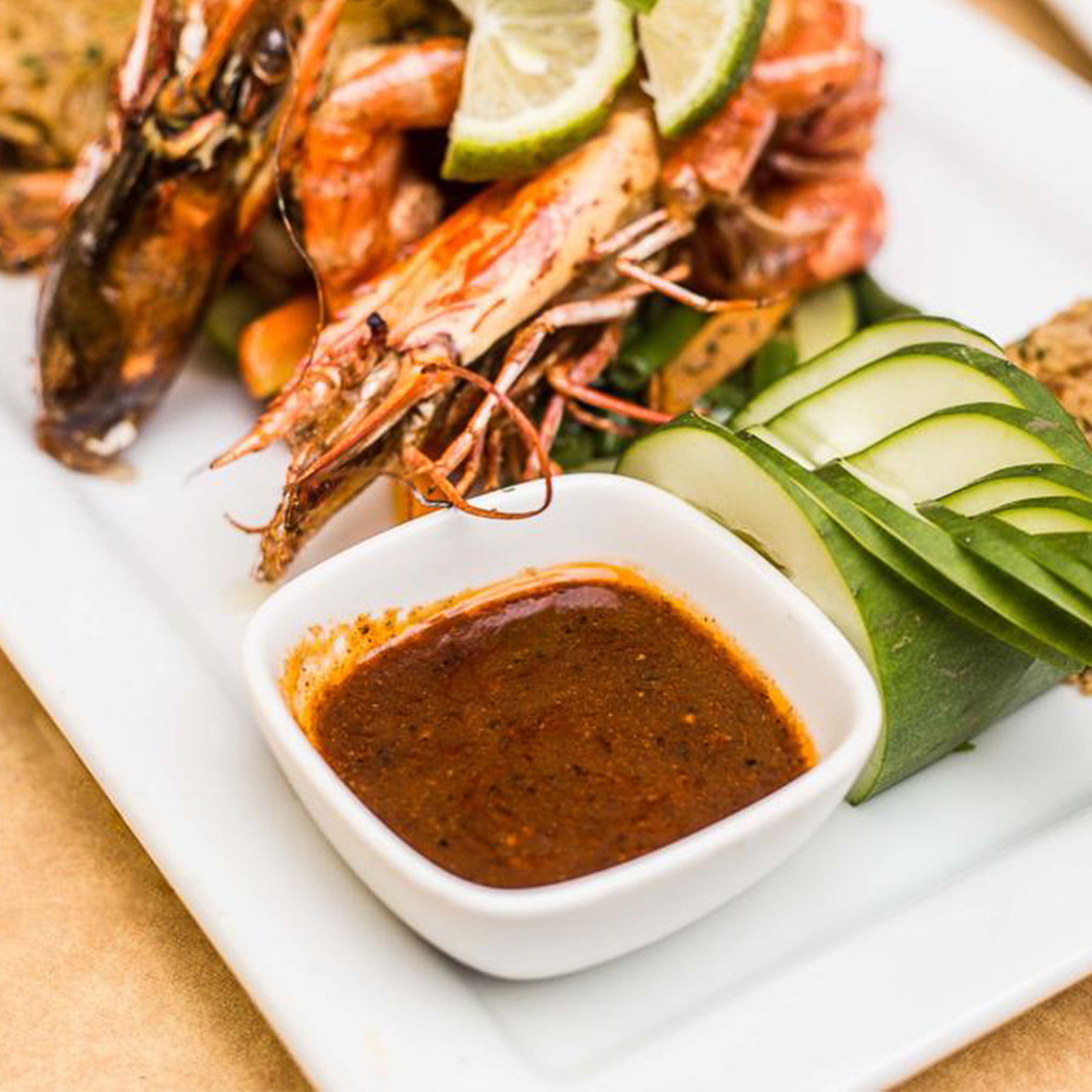 A plate of grilled shrimp with lime wedges and cucumber slices, accompanied by a small bowl of dipping sauce.