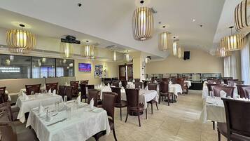 A spacious dining area with multiple tables and chairs, decorative lighting, and a television screen on the wall.