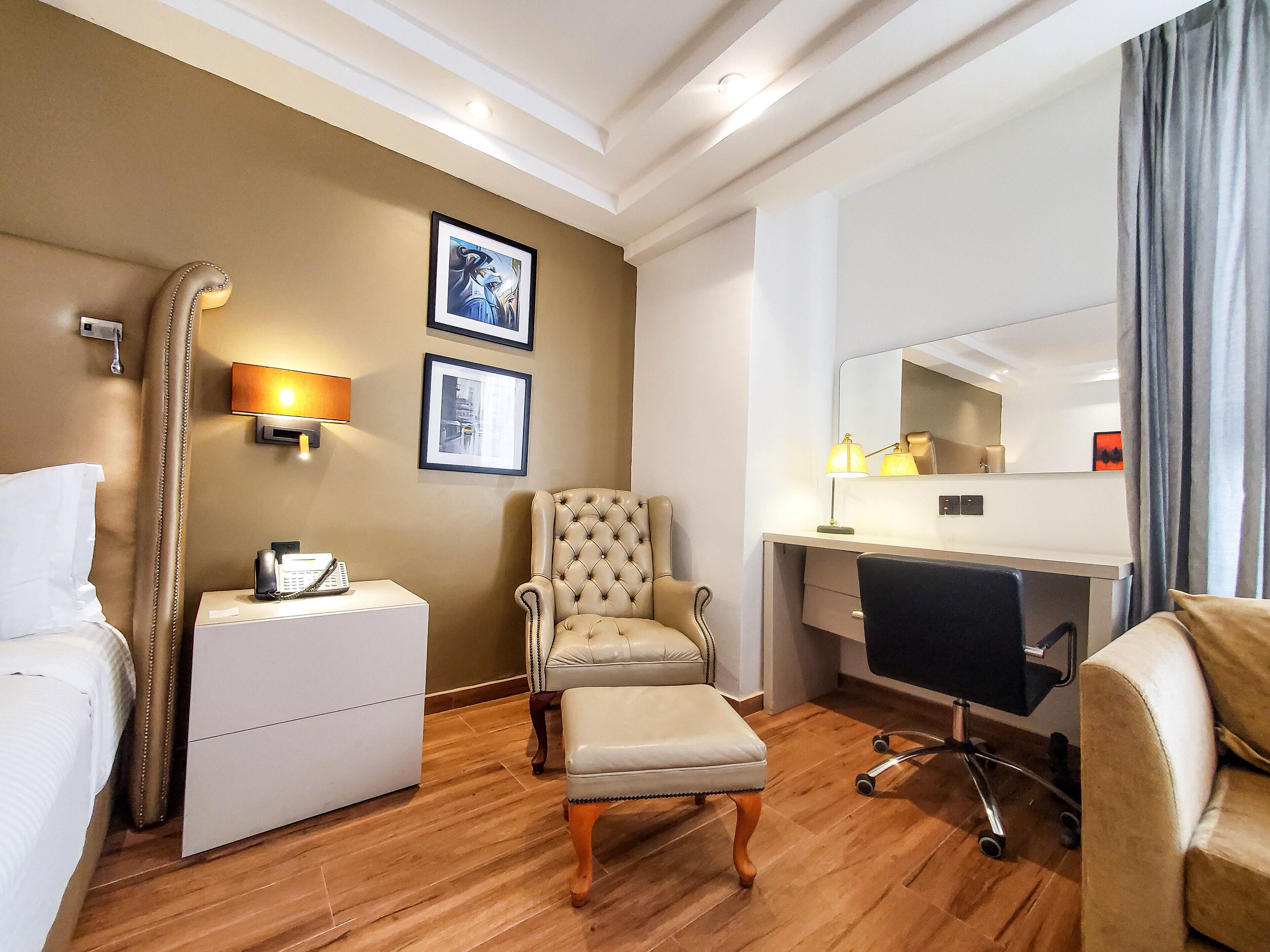 A hotel room with a bed, a desk, a chair, a small table, a telephone, and a wall-mounted lamp.