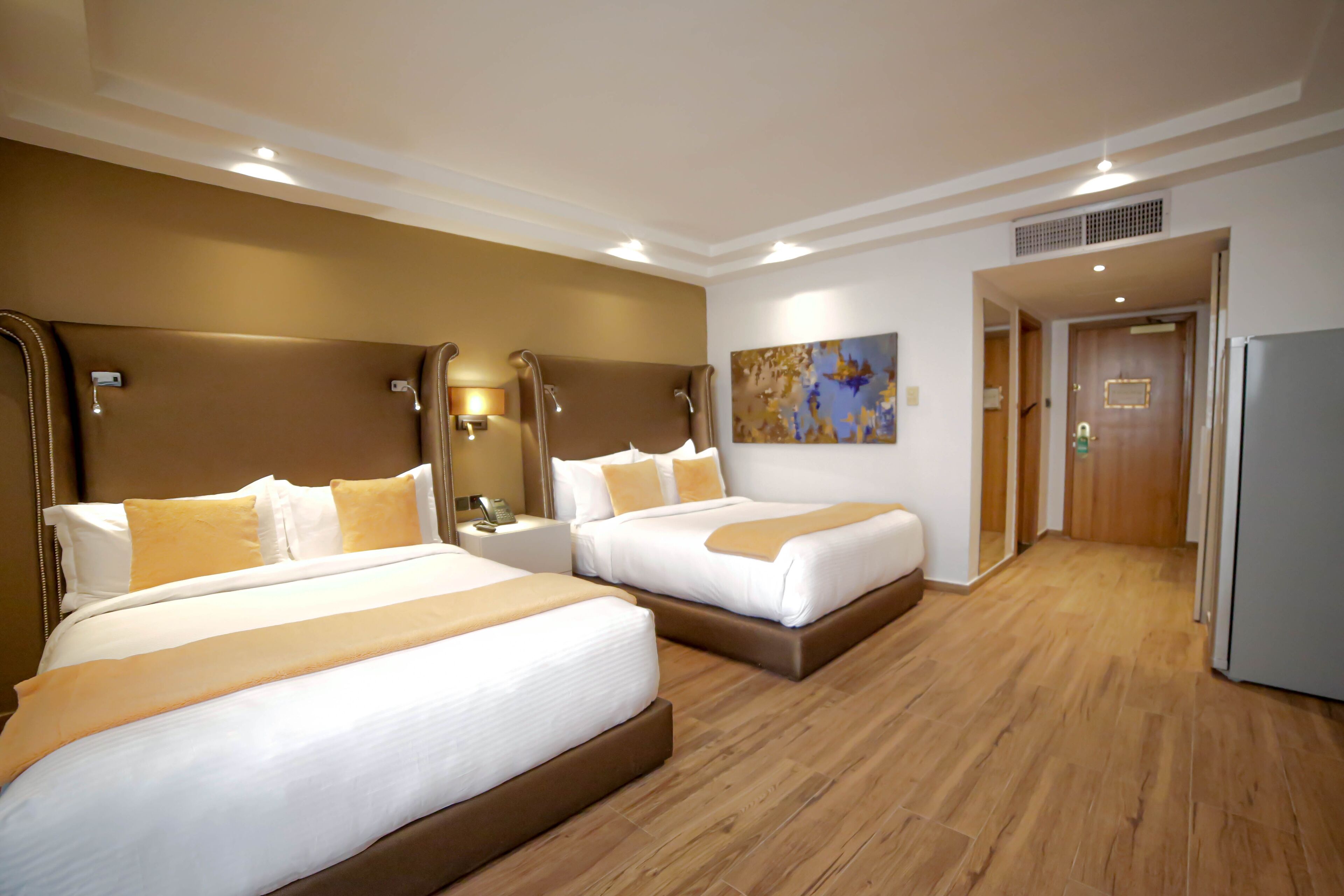 A hotel room with two beds, a wooden floor, a painting on the wall, and a door leading to another room.