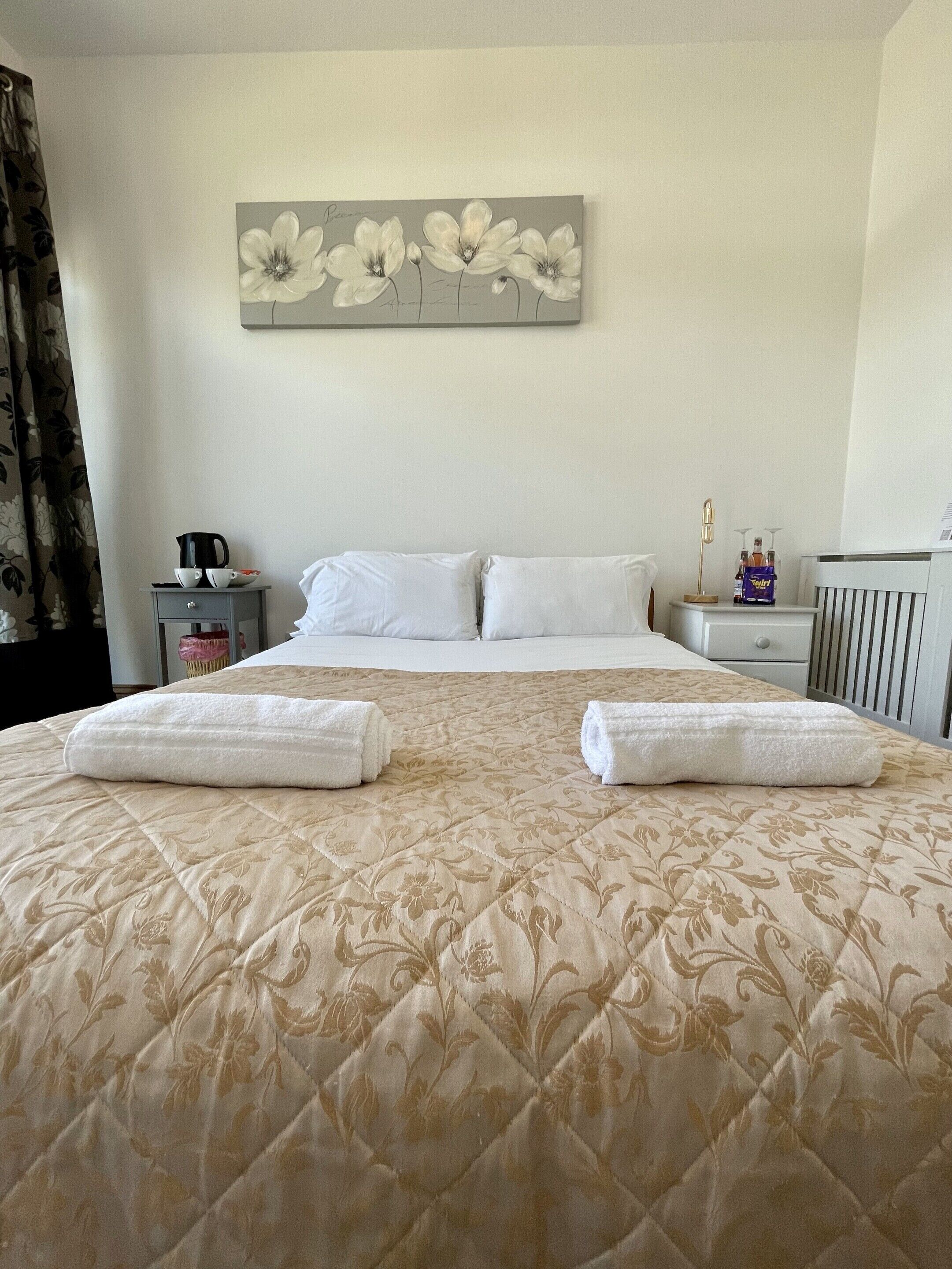 Un letto rifatto con cura, con una coperta a motivi floreali, due cuscini e due asciugamani piegati. È presente un comodino con una macchina per caffè e un piccolo oggetto decorativo. Sopra il letto, c'è un quadro con dei fiori.