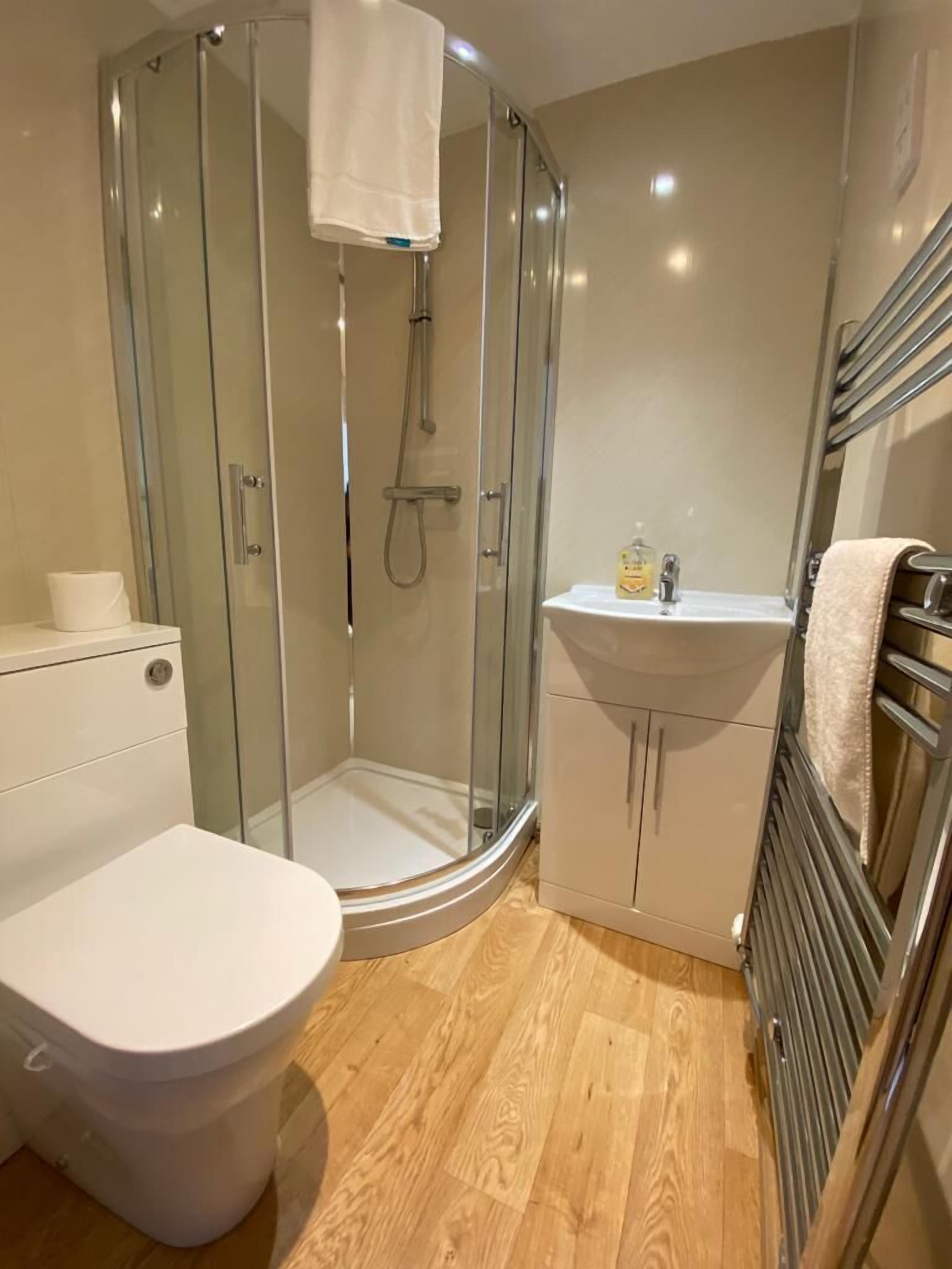 Un baño moderno con ducha envuelta en vidrio, inodoro blanco, lavamanos con espejo y perchero para toallas.