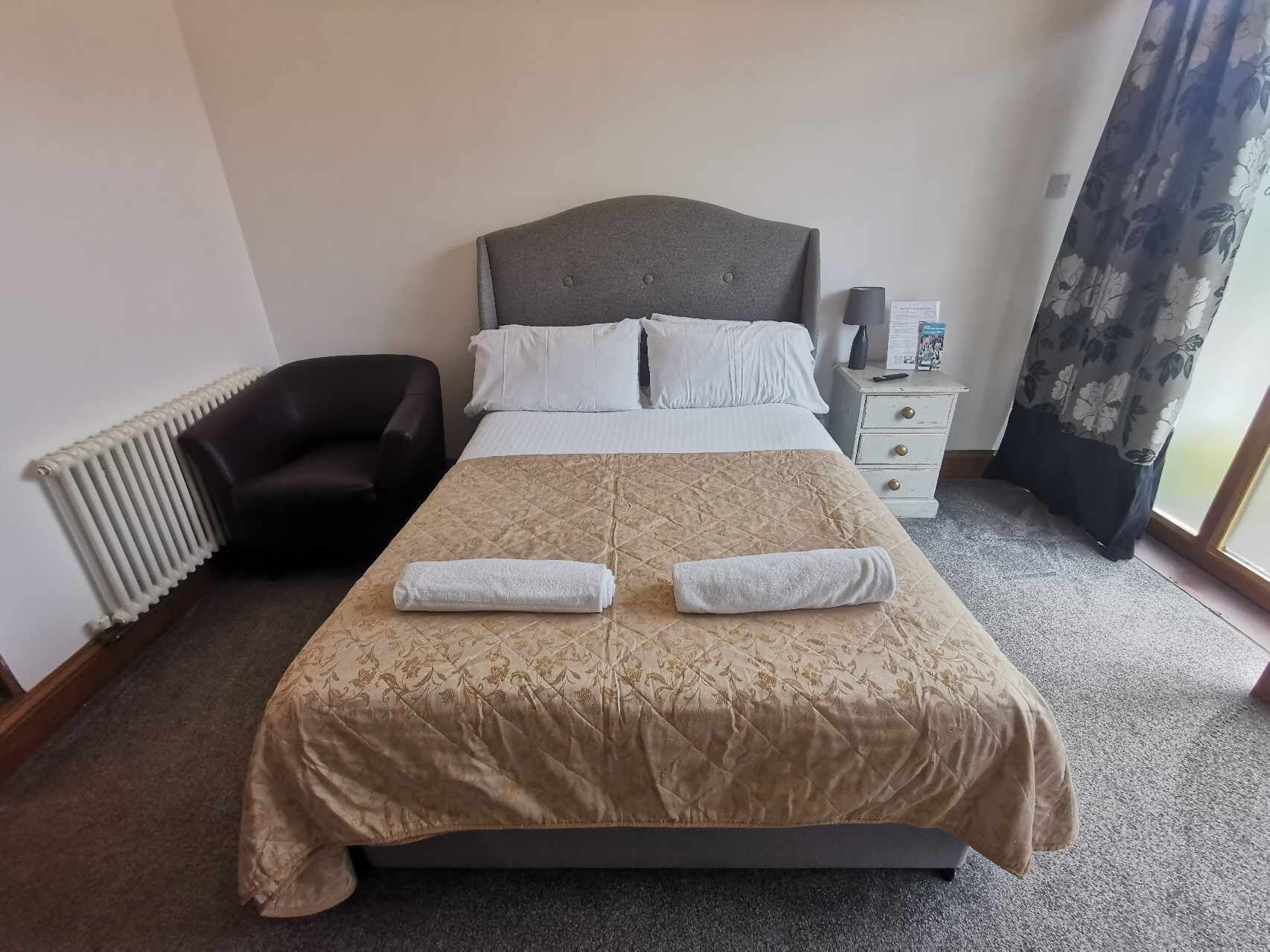 Una cama bien tendida con una colcha color beige, dos almohadas y dos toallas dobladas. A la izquierda, un sillón oscuro, una mesita de noche blanca con una lámpara y un radiador en la pared izquierda.