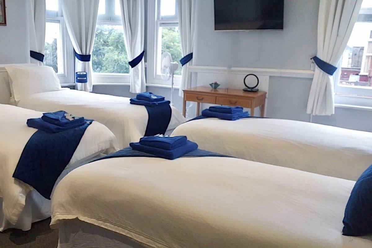 Habitación de hotel con dos camas, ropa de cama blanca, almohadas azules, una pequeña mesa de madera con un espejo y un televisor montado en la pared.