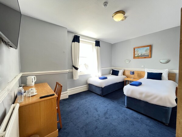 A hotel room with two beds, a desk, a chair, a window with curtains, a painting on the wall, and a ceiling light. - Royal Hotel Sheerness (Sheerness)