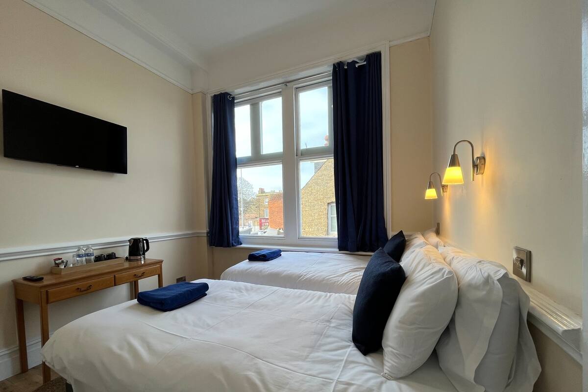 Habitación de hotel con dos camas, un televisor de pantalla plana montado en la pared, una mesita de noche con hervidor y tazas, y una ventana con cortinas.