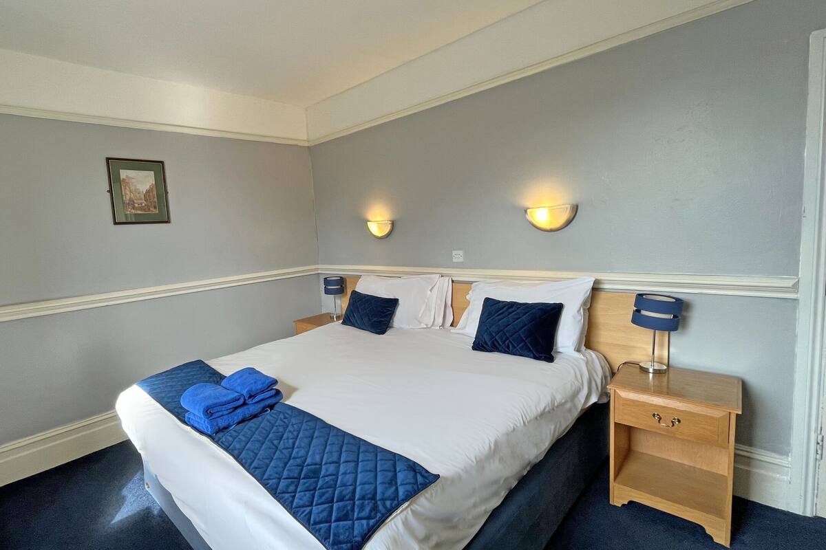 Una cama bien hecha con colcha y almohadas azules, una mesita de noche con lámpara, y una lámpara fijada a la pared.