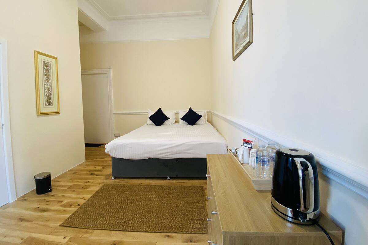 Habitación de hotel con cama, mesita de noche con tetera, cubo de basura pequeño y un cuadro en la pared.