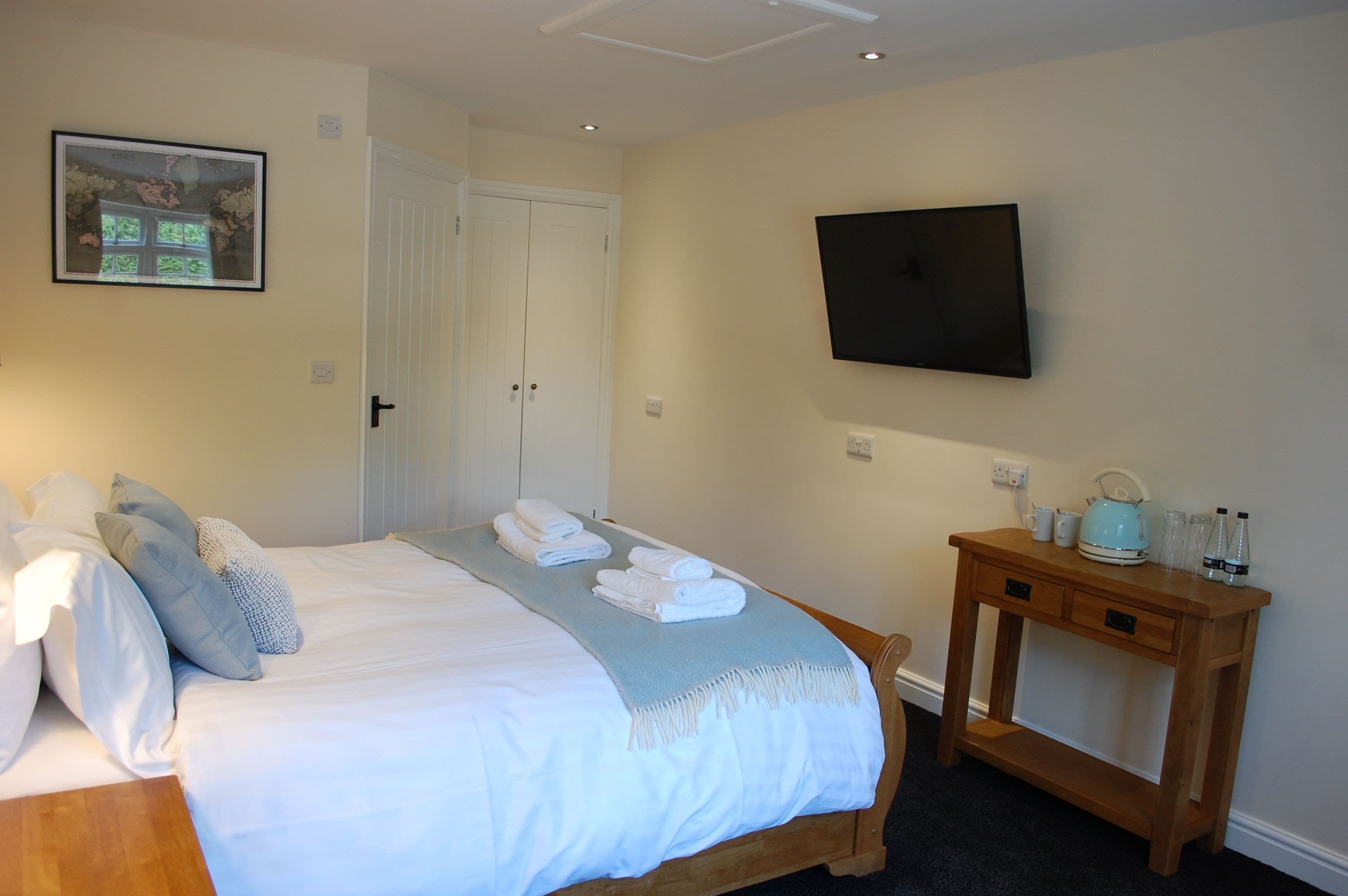 A neatly made bed with white linens and a blue throw, a wooden bedside table with a kettle and glasses, and a flat-screen TV mounted on the wall.