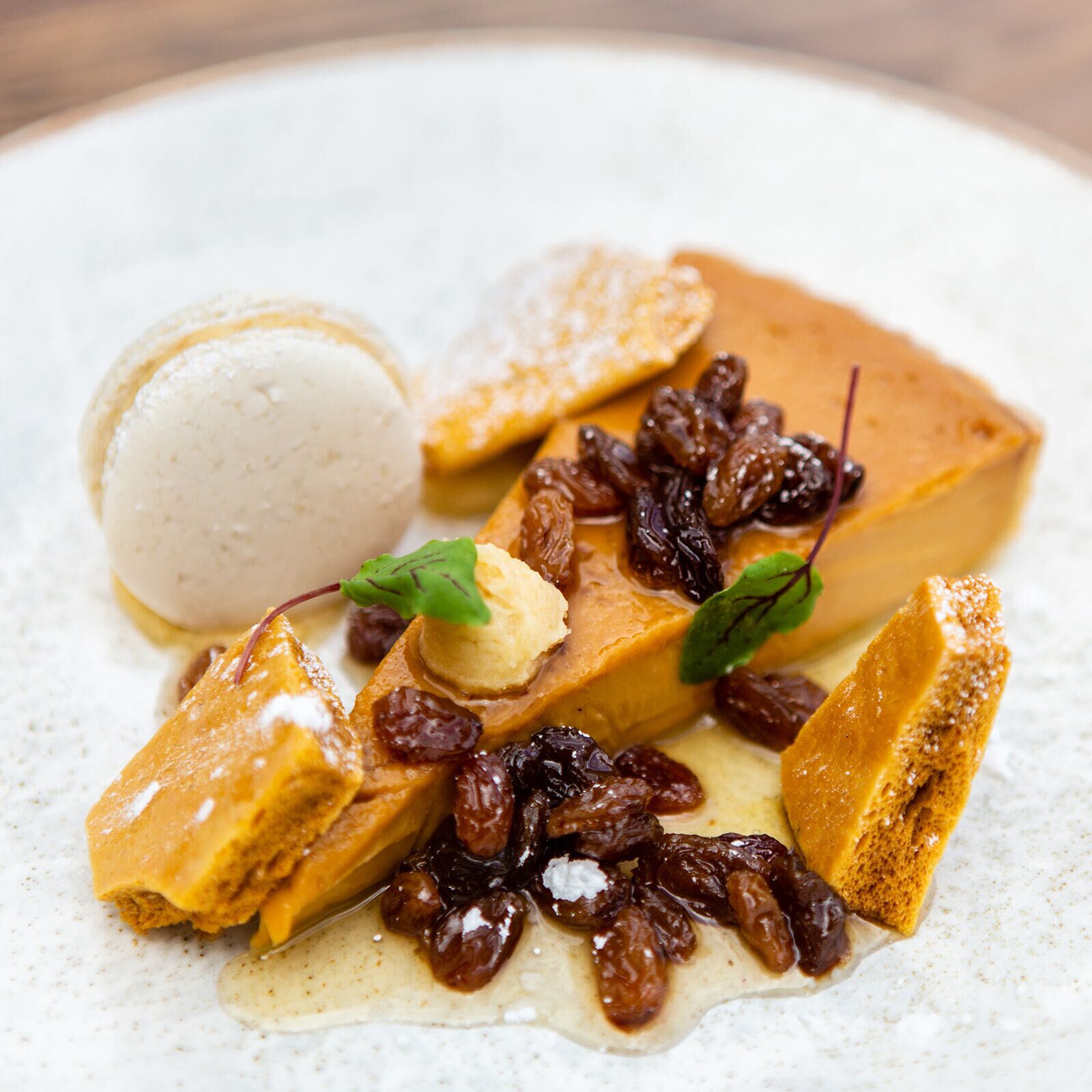 Une assiette de dessert comprenant un flan au caramel, une boule de glace à la vanille et une tranche de pain garnie de caramel et de raisins secs.
