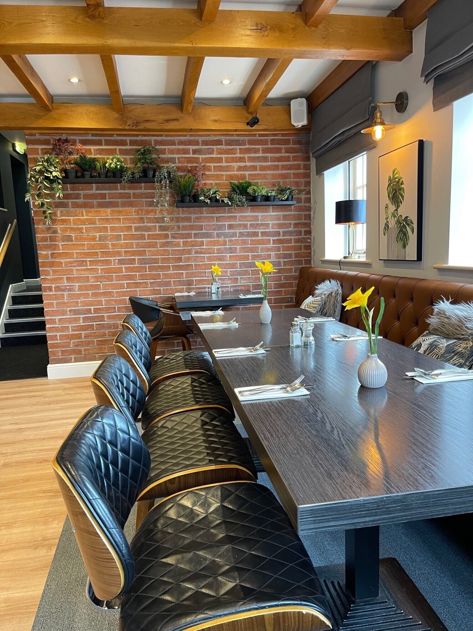 A long dining table with a bench seating arrangement, a brick wall with greenery, and a window with a plant.