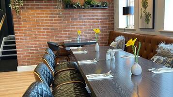 A long dining table with a bench seating arrangement, a brick wall with greenery, and a window with a plant.