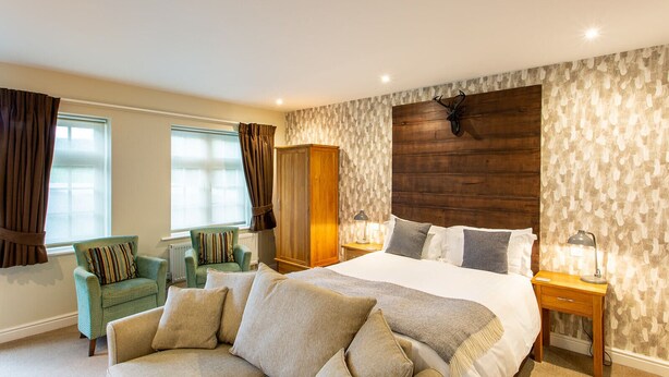 A spacious bedroom with a large bed, a wooden headboard, a green armchair, a wooden wardrobe, and a window with curtains. - The Royal Oak - Inn (Loughborough)