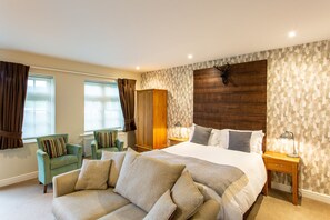 Une chambre spacieuse avec un grand lit, une tête de lit en bois, un fauteuil vert, une armoire en bois et une fenêtre avec des rideaux.