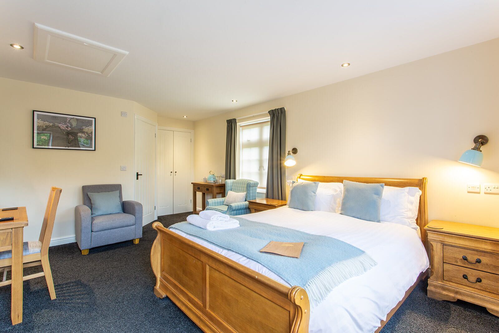A bedroom with a wooden bed, a grey armchair, a small table, and a desk with a chair.