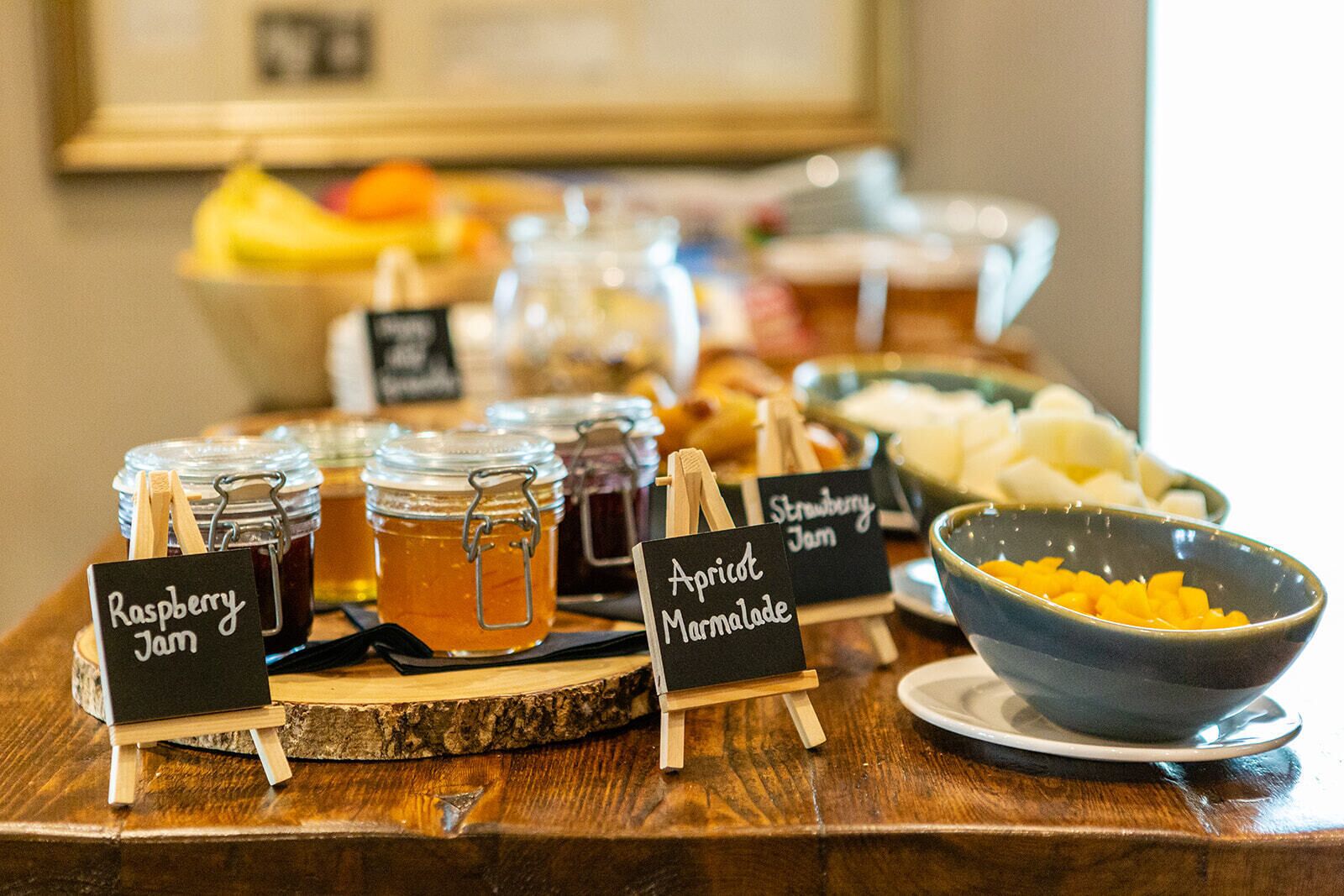 A buffet table with jars of raspberry jam, apricot marmalade, and strawberry jam, along with bowls of fruit.