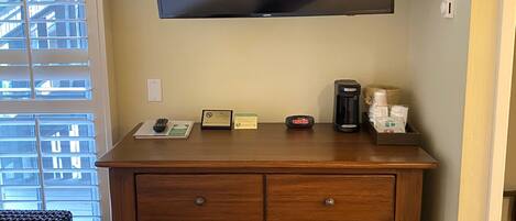 A hotel room with a large flat-screen TV mounted on the wall, a wooden dresser with drawers, a small table with a clock and a framed certificate, and a window with blinds.