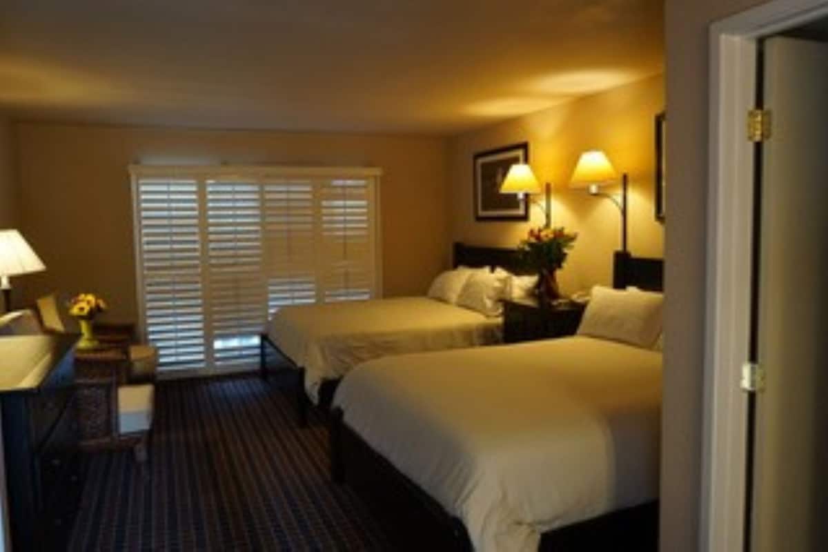 A hotel room with two beds, a desk with a lamp, and a window with blinds.