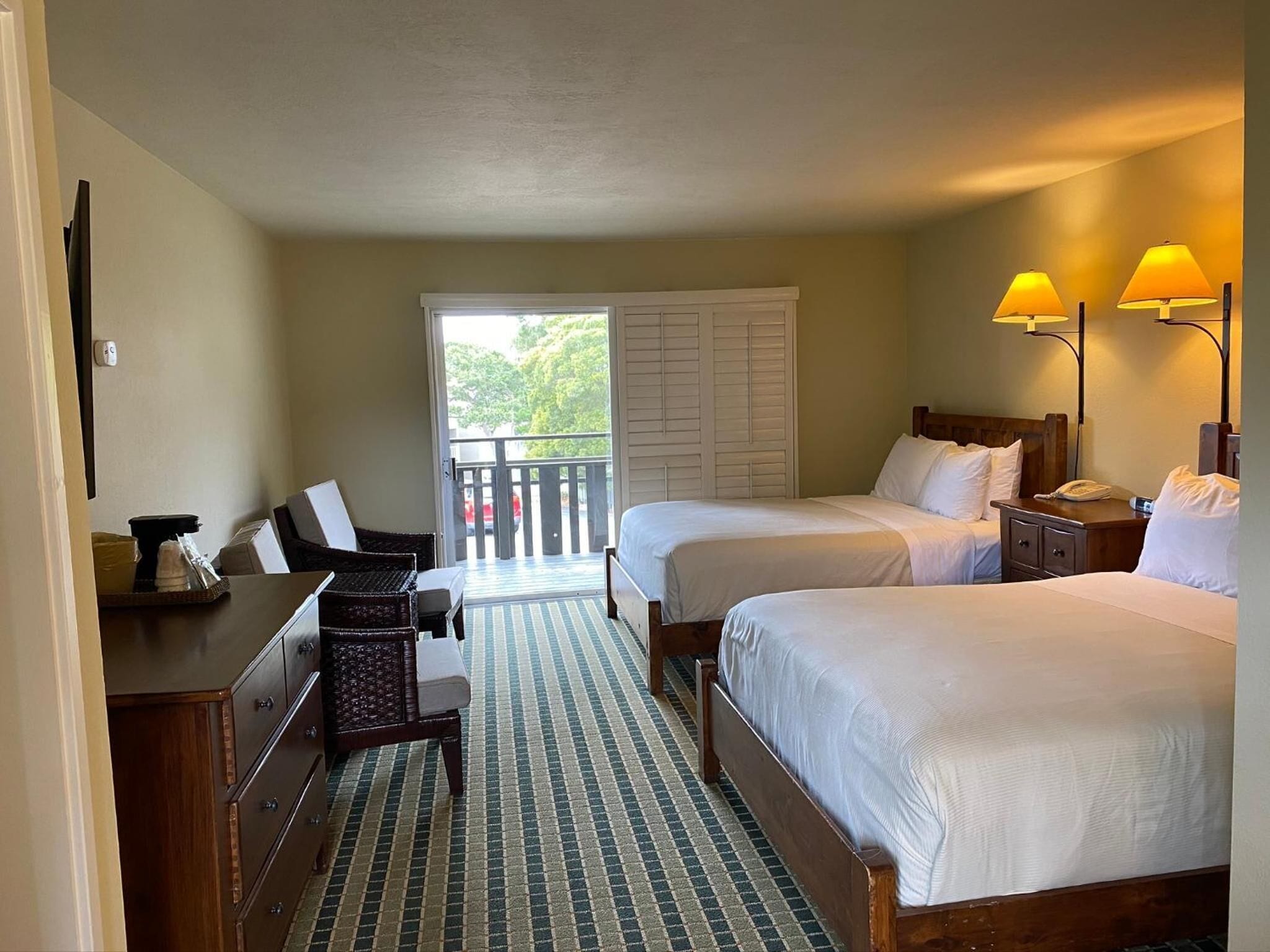 A hotel room with two beds, a desk, a chair, and a balcony with a view of trees.