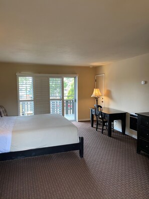 A hotel room with a large bed, a desk with a chair, a lamp, and a view through the window. - Olympia Lodge (Pacific Grove)