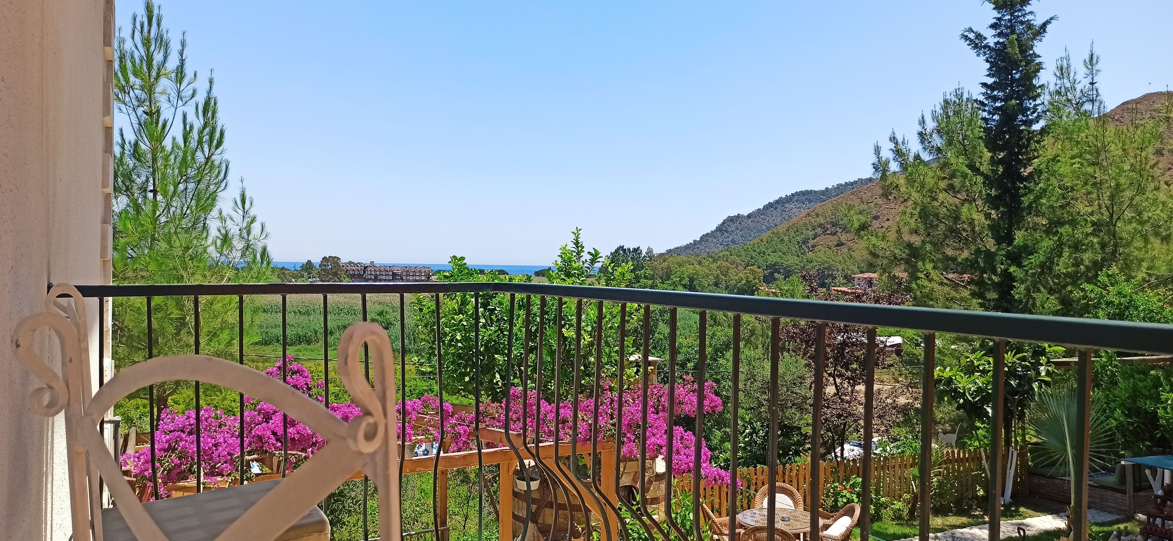 A balcony with a view of a garden, a wooden fence, and a distant body of water.
