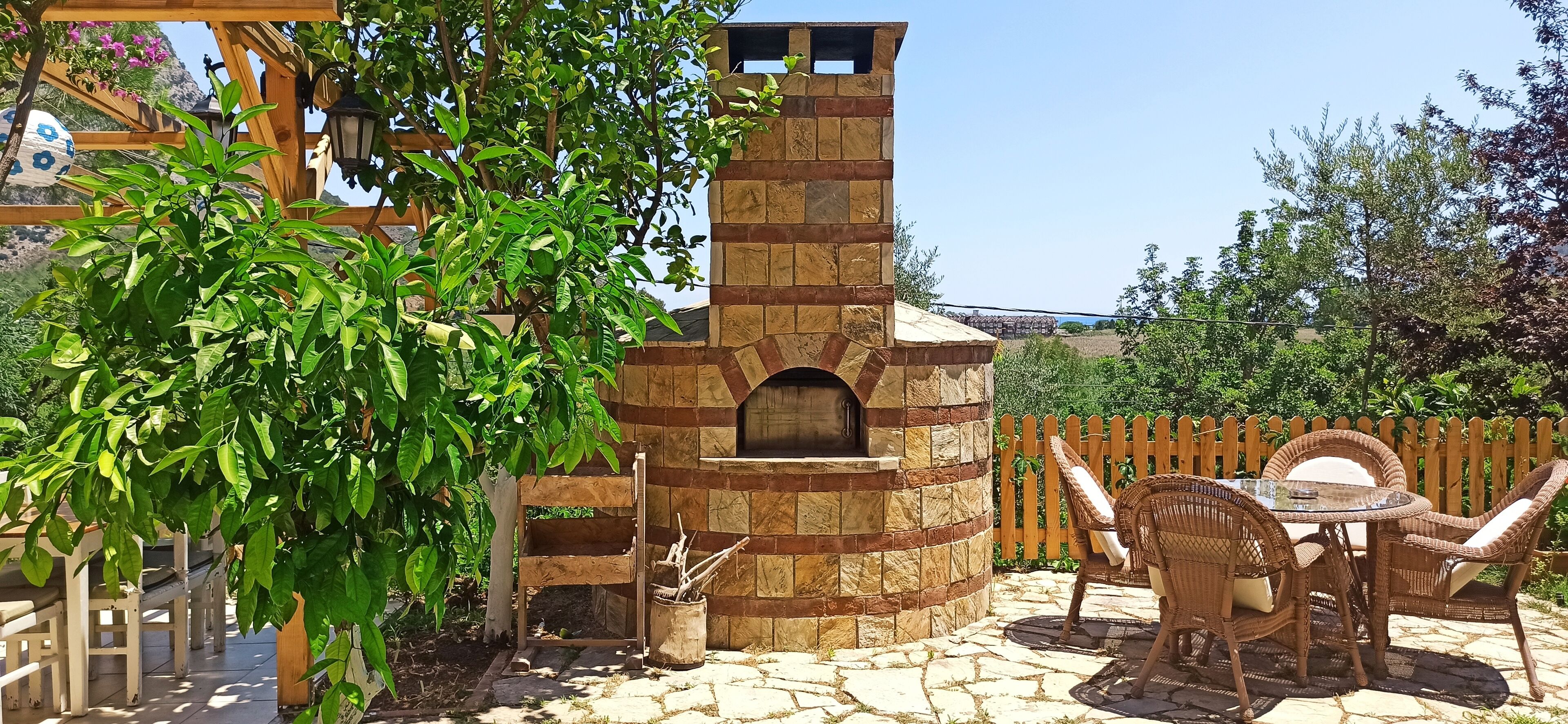 A stone-built outdoor kitchen with a pizza oven, wicker furniture, and a wooden pergola.