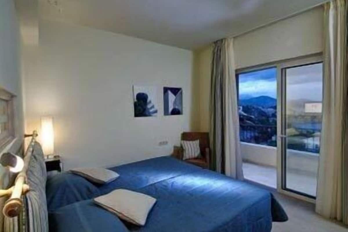 Habitación de hotel con cama, mesita de noche, lámpara, ventana con vistas y dos cuadros colgados.