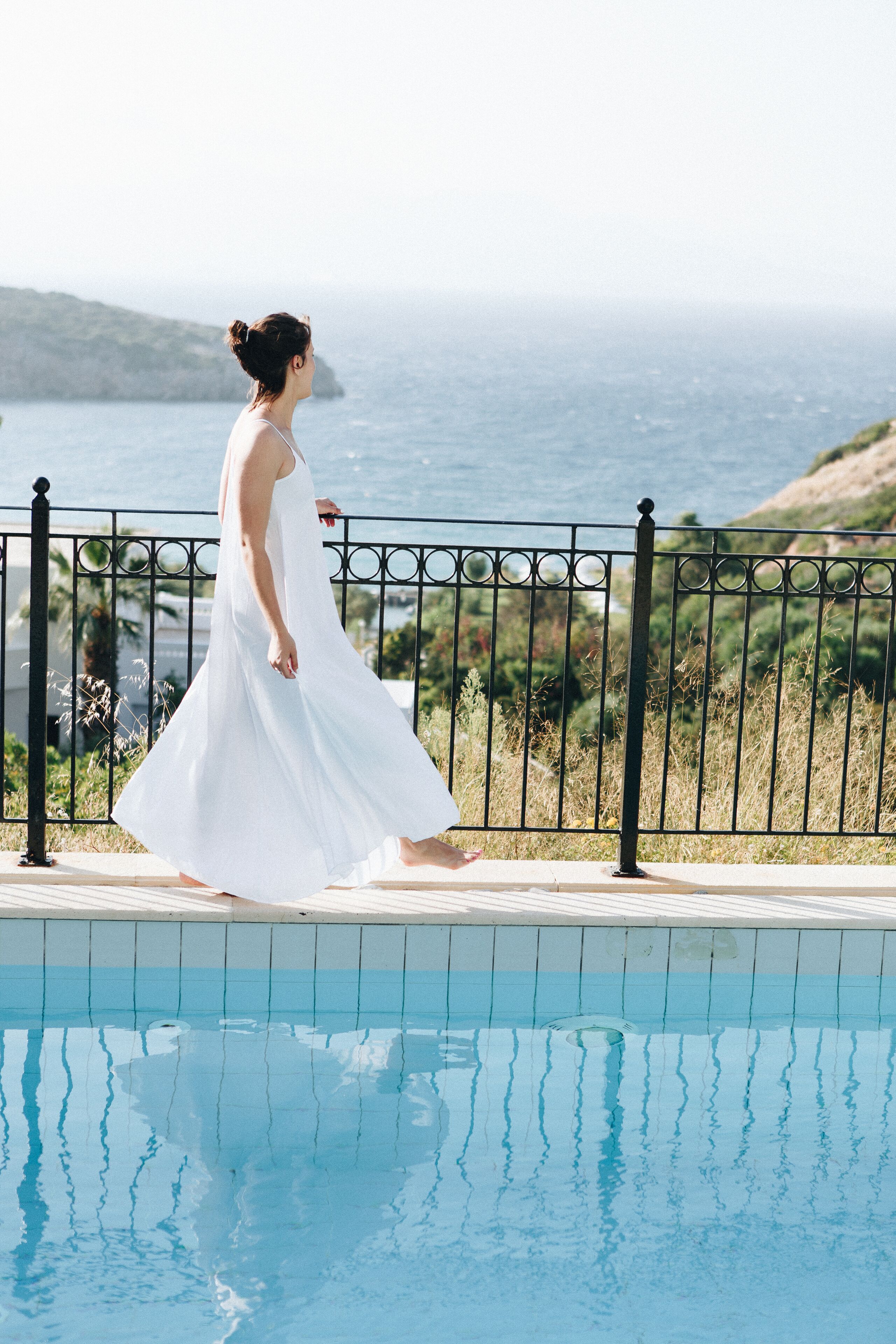 Une personne vêtue d’une robe blanche se promène au bord d’une piscine, avec vue sur la mer et les collines.