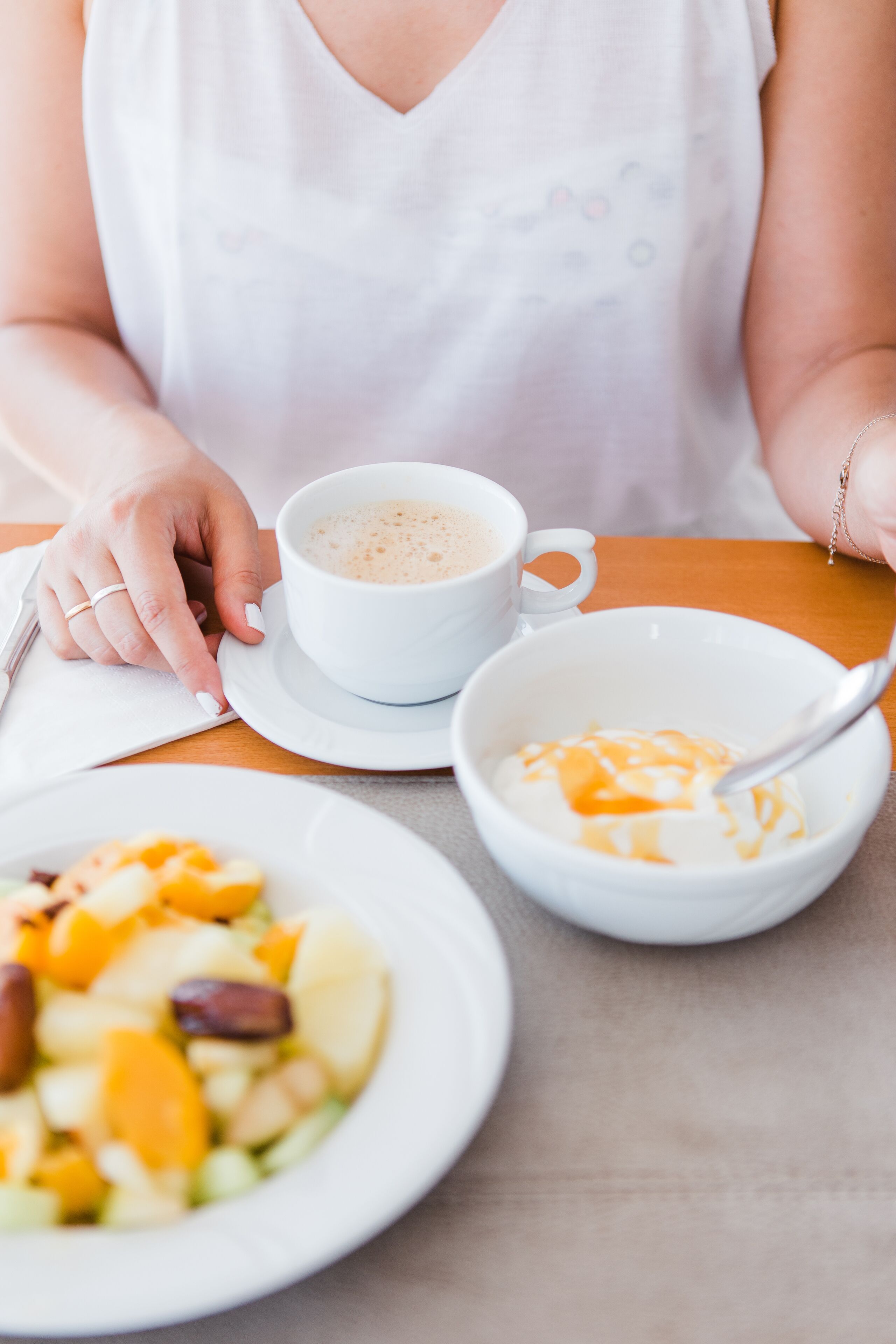 A person sitting at a table with a cup of coffee, a bowl of yogurt, and a plate of fruit salad.