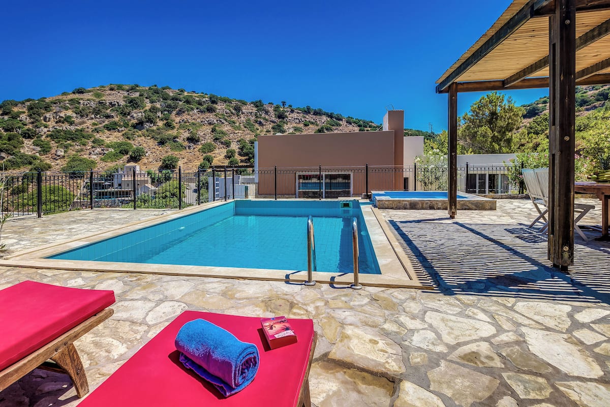 Una piscina con suelo de piedra, sillas de descanso rojas y una estructura de pérgola de madera.