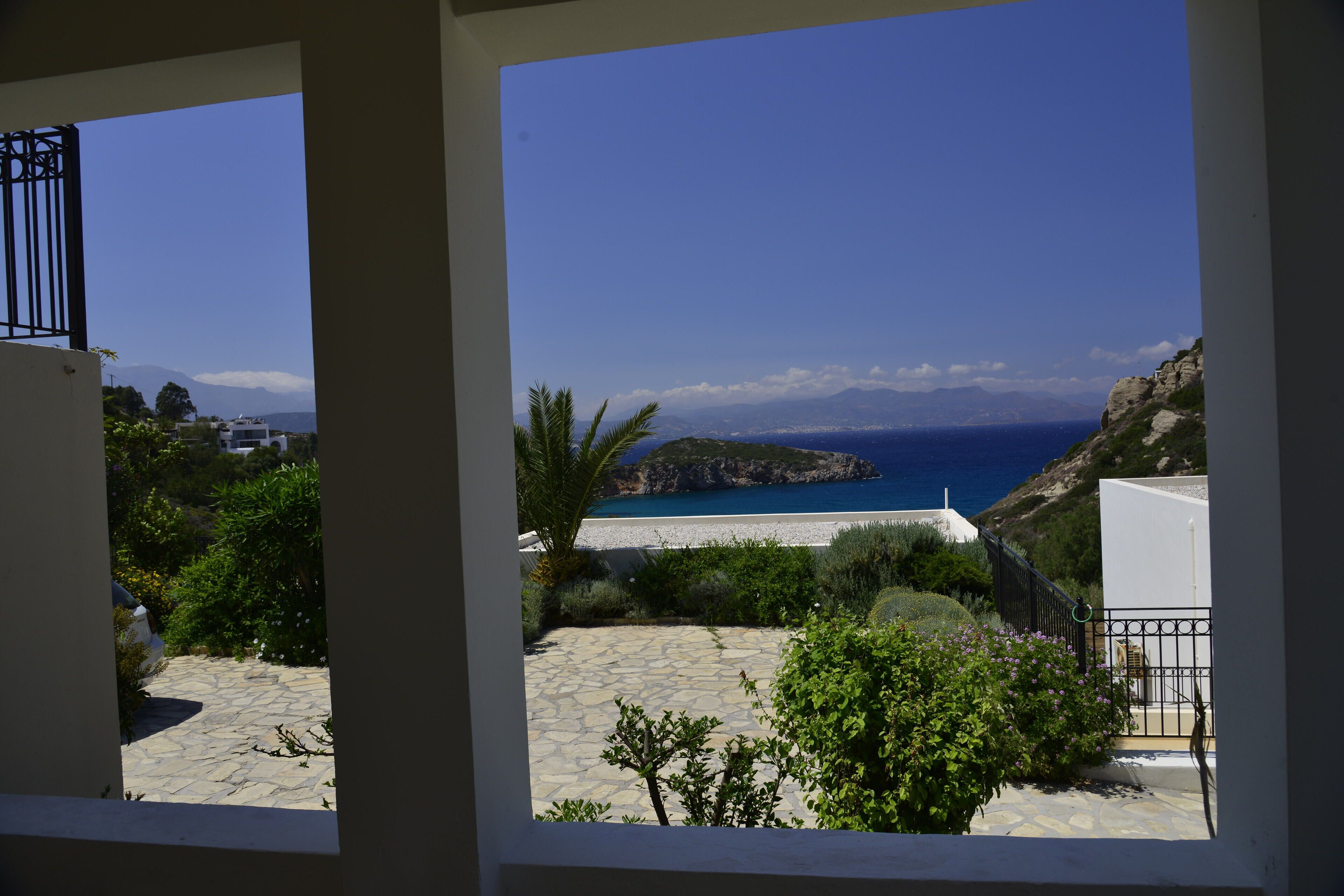 Blick durch ein Fenster auf einen steingepflasterten Innenhof mit einem weißen Gebäude, einem schwarzen Metallgitter und einer üppigen Grünfläche, die zu einem Strand mit Meer und Bergen im Hintergrund führt.