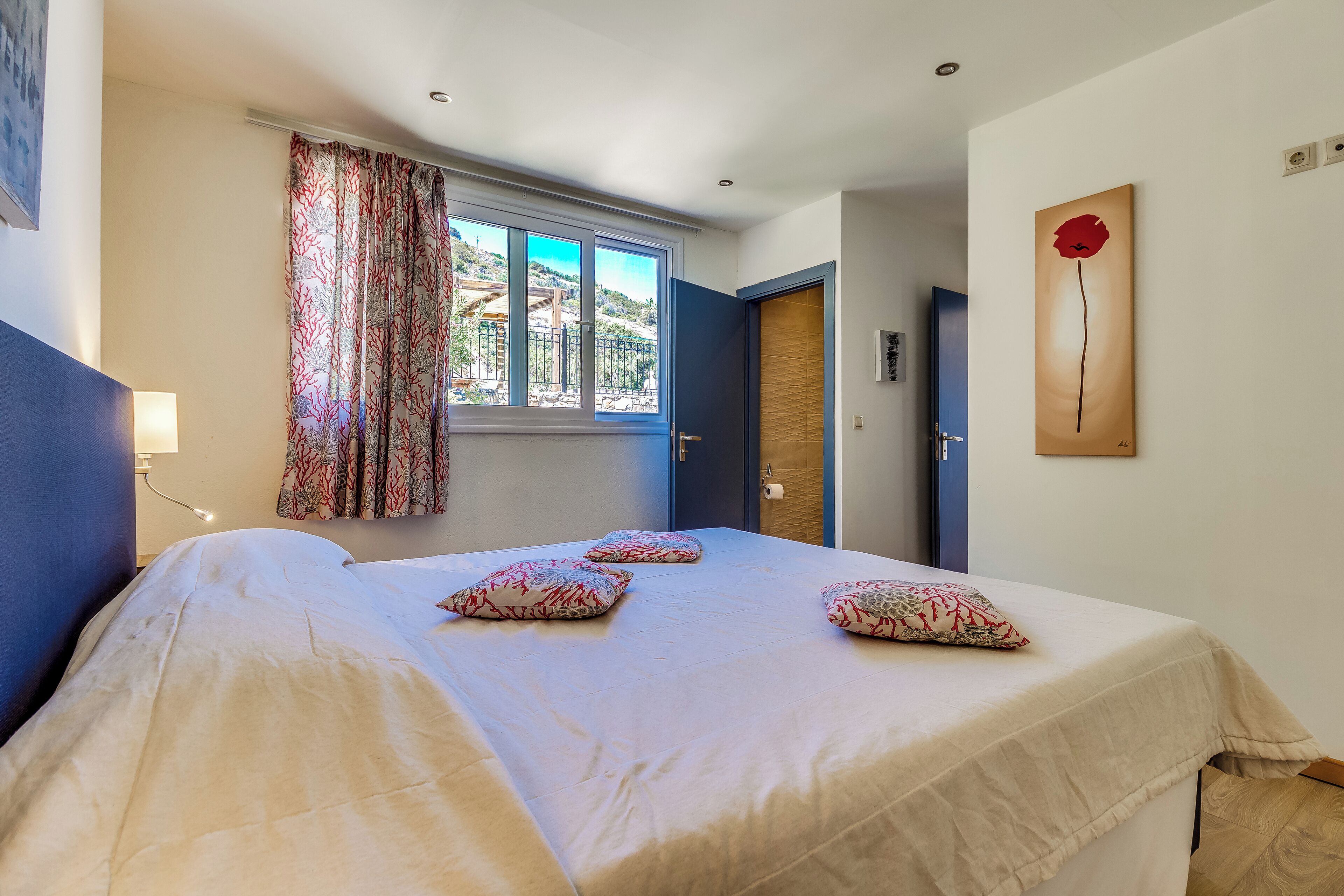 Un dormitorio con cama, almohadas estampadas, ventana con cortinas, puerta y un cuadro en la pared.
