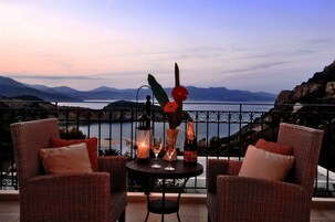 Un conjunto de sillón y mesa de mimbre con una botella, vasos y una vela en un balcón con vistas a un cuerpo de agua y montañas.