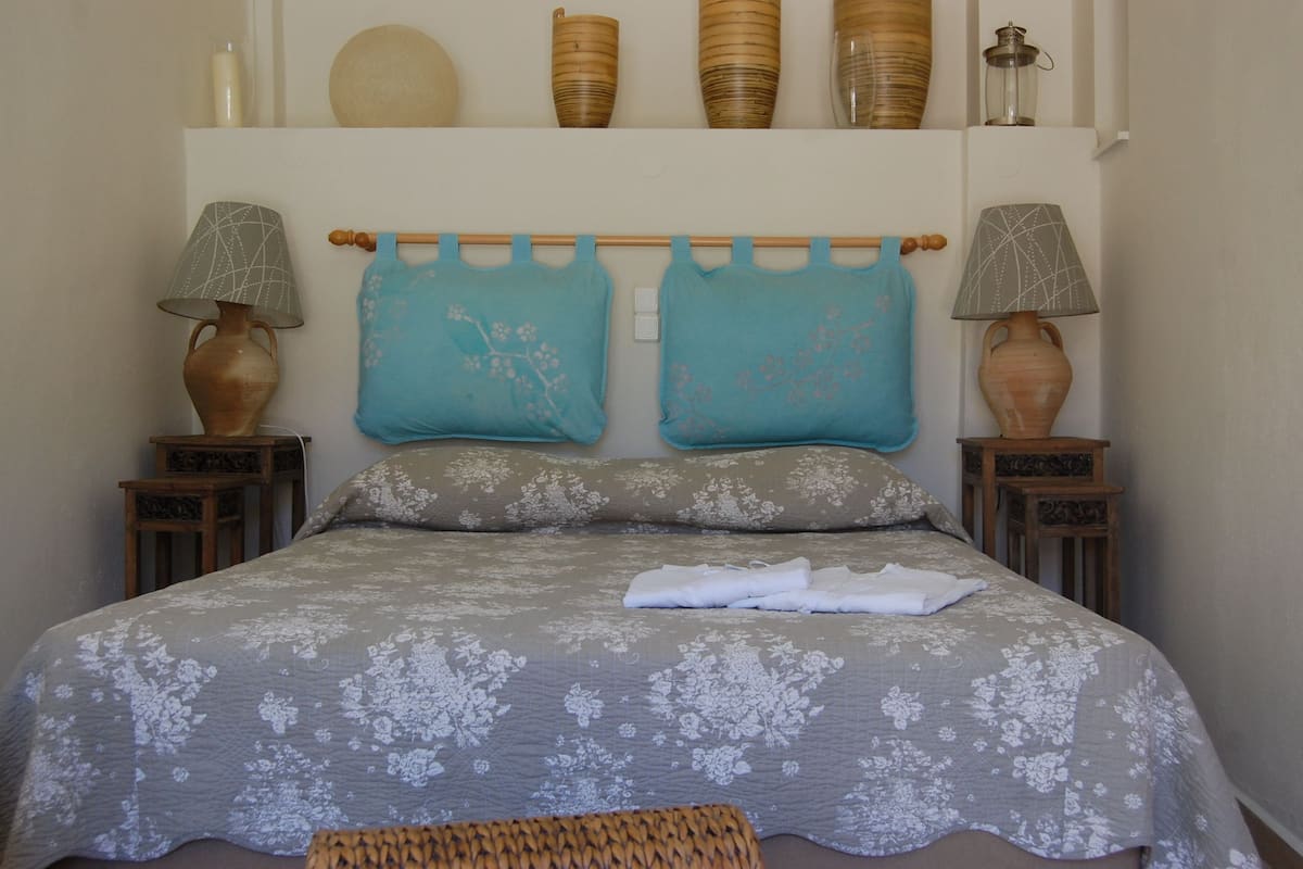 Una cama bien hecha con ropa de cama floral, dos lámparas fijadas a la pared y objetos decorativos en un estante superior.