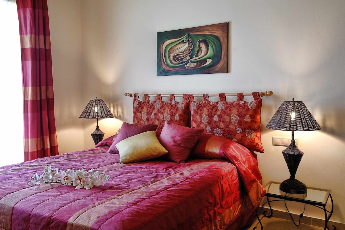 Una cama con una colcha roja, varios almohadones y un arreglo decorativo de orquídeas. Hay dos lámparas fijadas a la pared a cada lado de la cama.