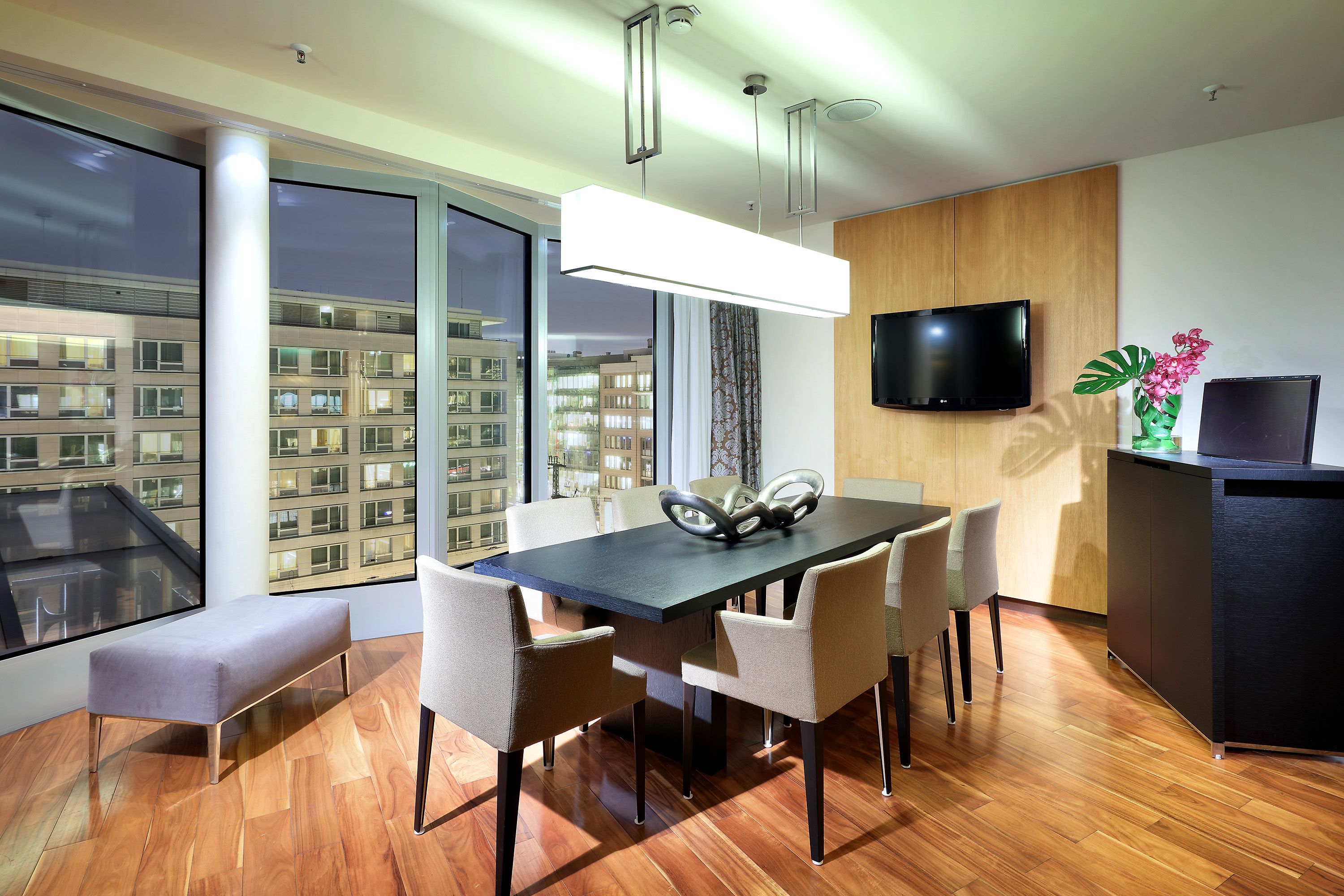 Un comedor moderno con una mesa rectangular, seis sillas, un banco, un televisor de pantalla plana montado en la pared y un ventanal con vista a la ciudad.