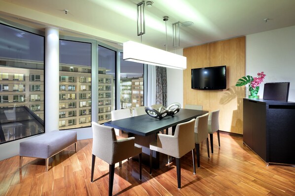 Un comedor moderno con una mesa rectangular, seis sillas, un banco, un televisor de pantalla plana montado en la pared y un ventanal con vista a la ciudad.