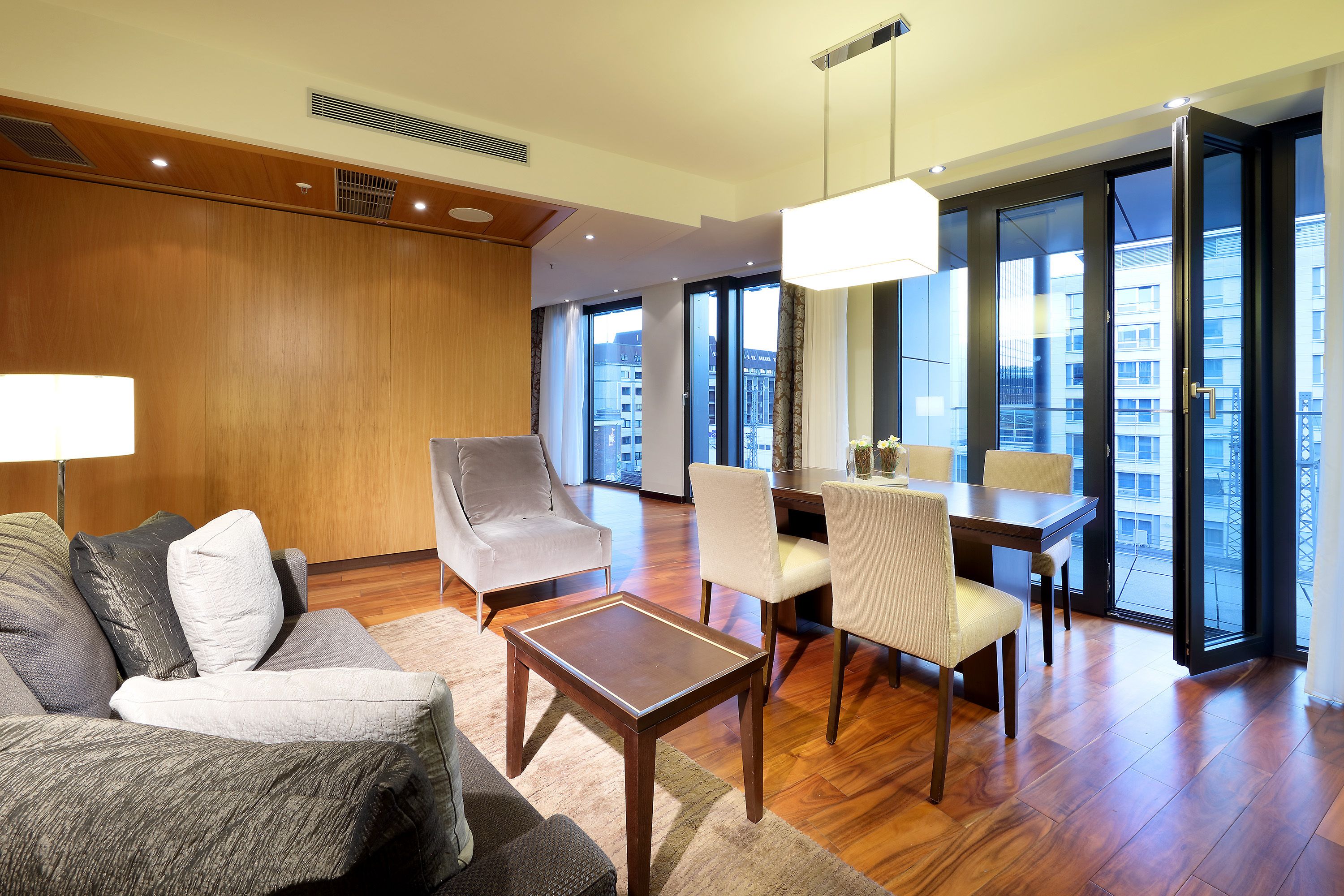 Ein modernes Hotelzimmer mit einer Wandpaneele aus Holz, einer Couch, einem Couchtisch, einem Essbereich mit Stühlen und großen Fenstern mit Blick auf die Stadt.