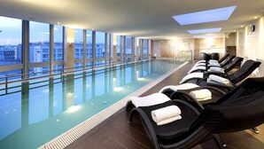 Une piscine intérieure moderne, dotée de chaises longues et d’une rambarde en verre, offrant une vue sur le paysage urbain.