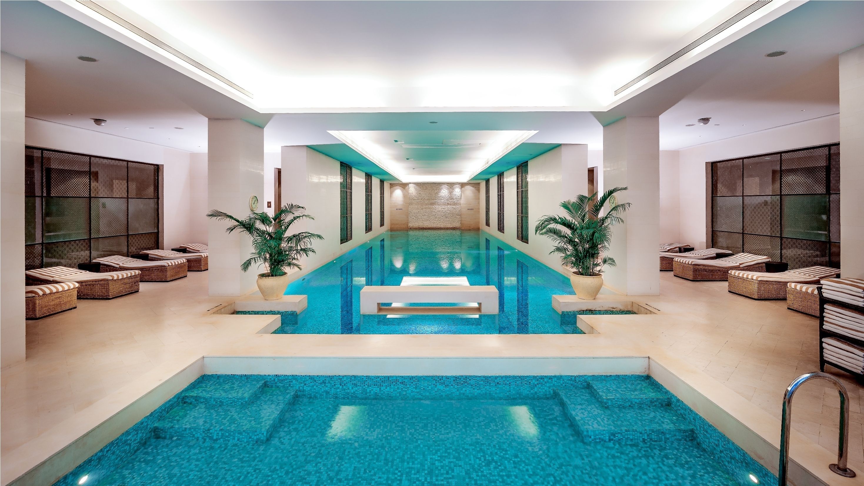 Una piscina interior moderna con agua azul cristalina, rodeada de zonas de descanso con mobiliario de mimbre y plantas en macetas.